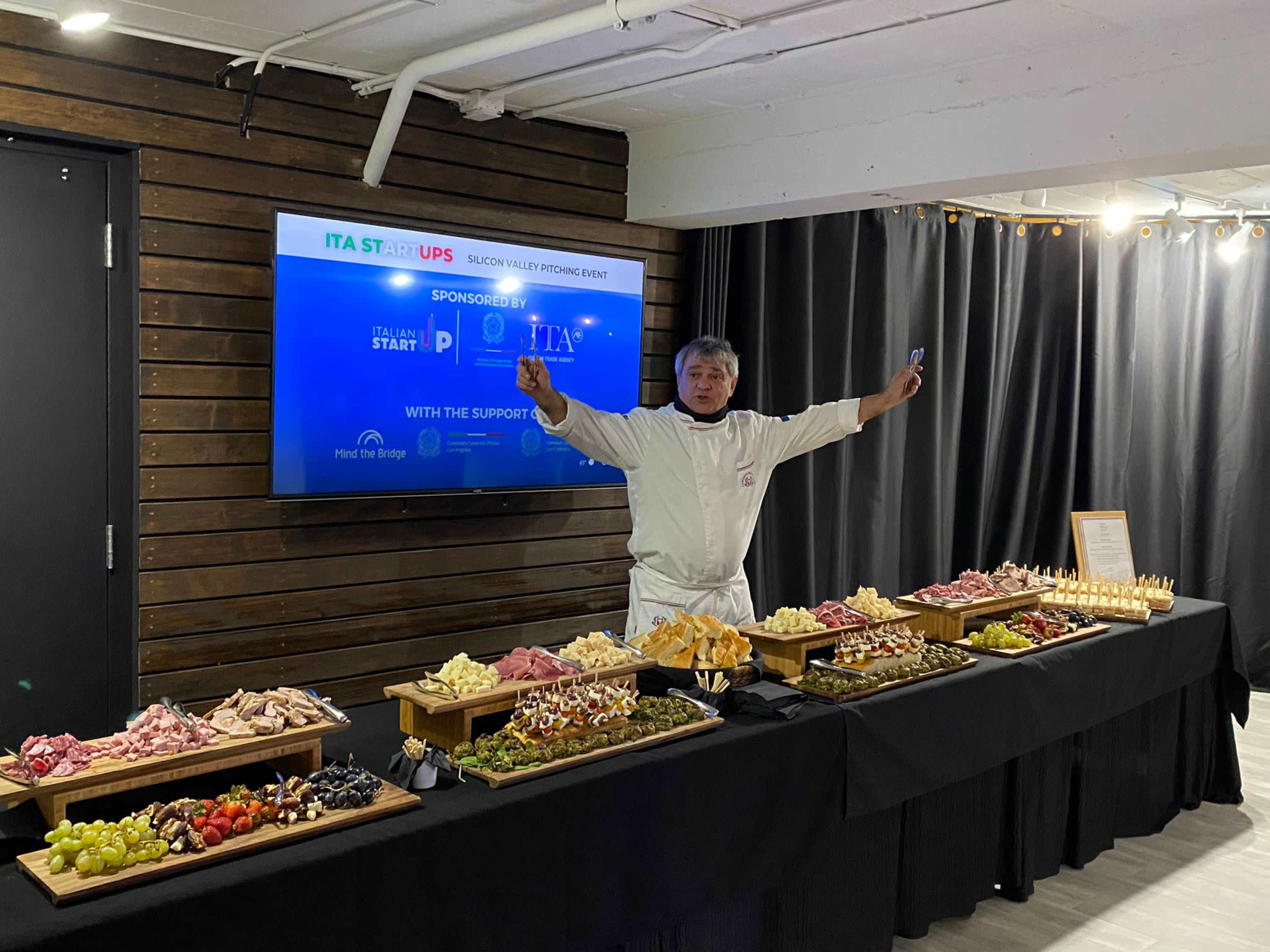 A chef gestures enthusiastically in front of a long table filled with various platters of charcuterie and appetizers at an event featuring signage for ITA Startups.