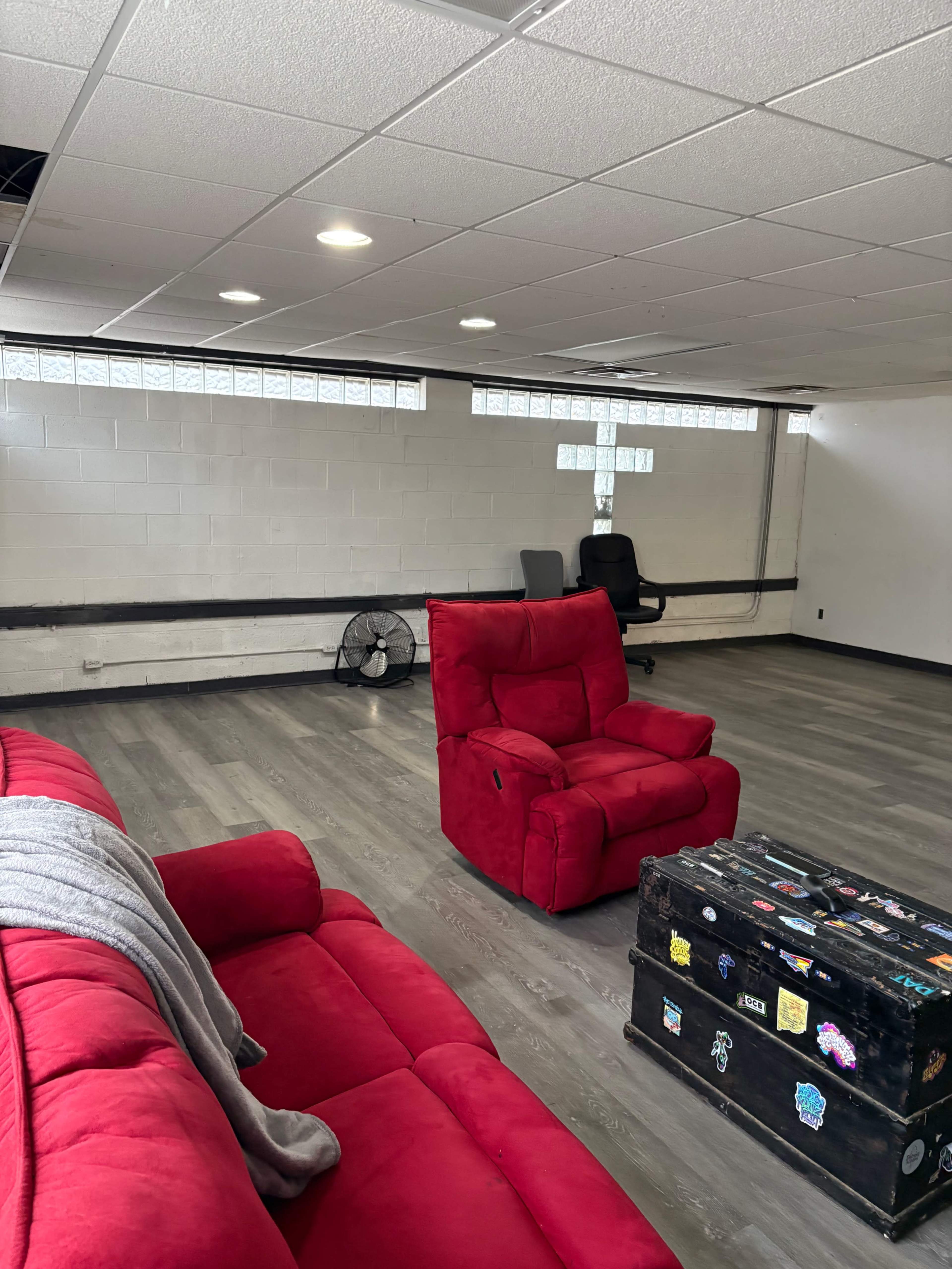 The image shows a spacious room with red recliner sofas, a fan, and a wooden trunk, featuring a few chairs and natural light from high windows.