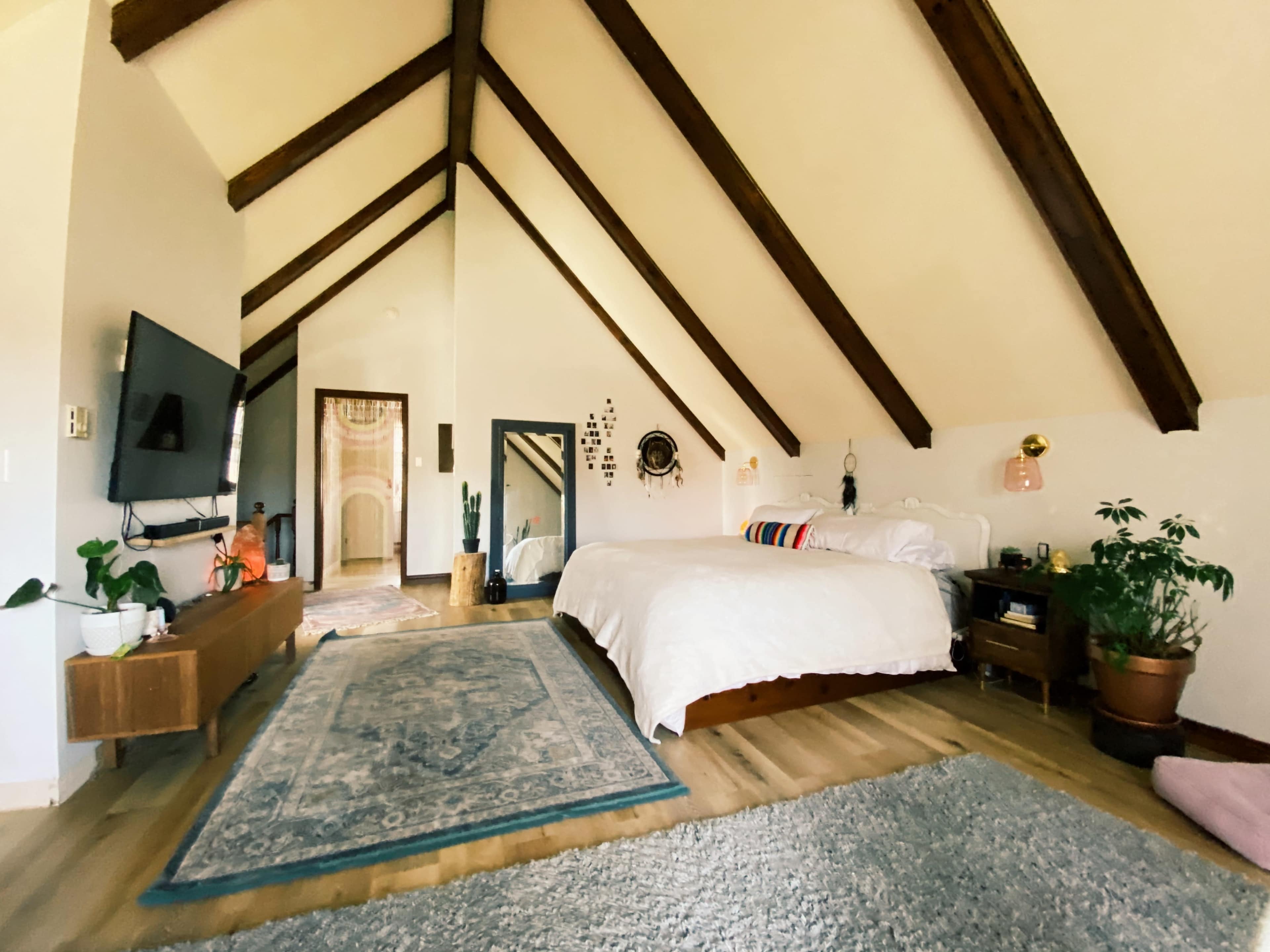 A spacious bedroom with a high, vaulted ceiling, featuring a large bed, a television mounted on the wall, and plants placed around the room.