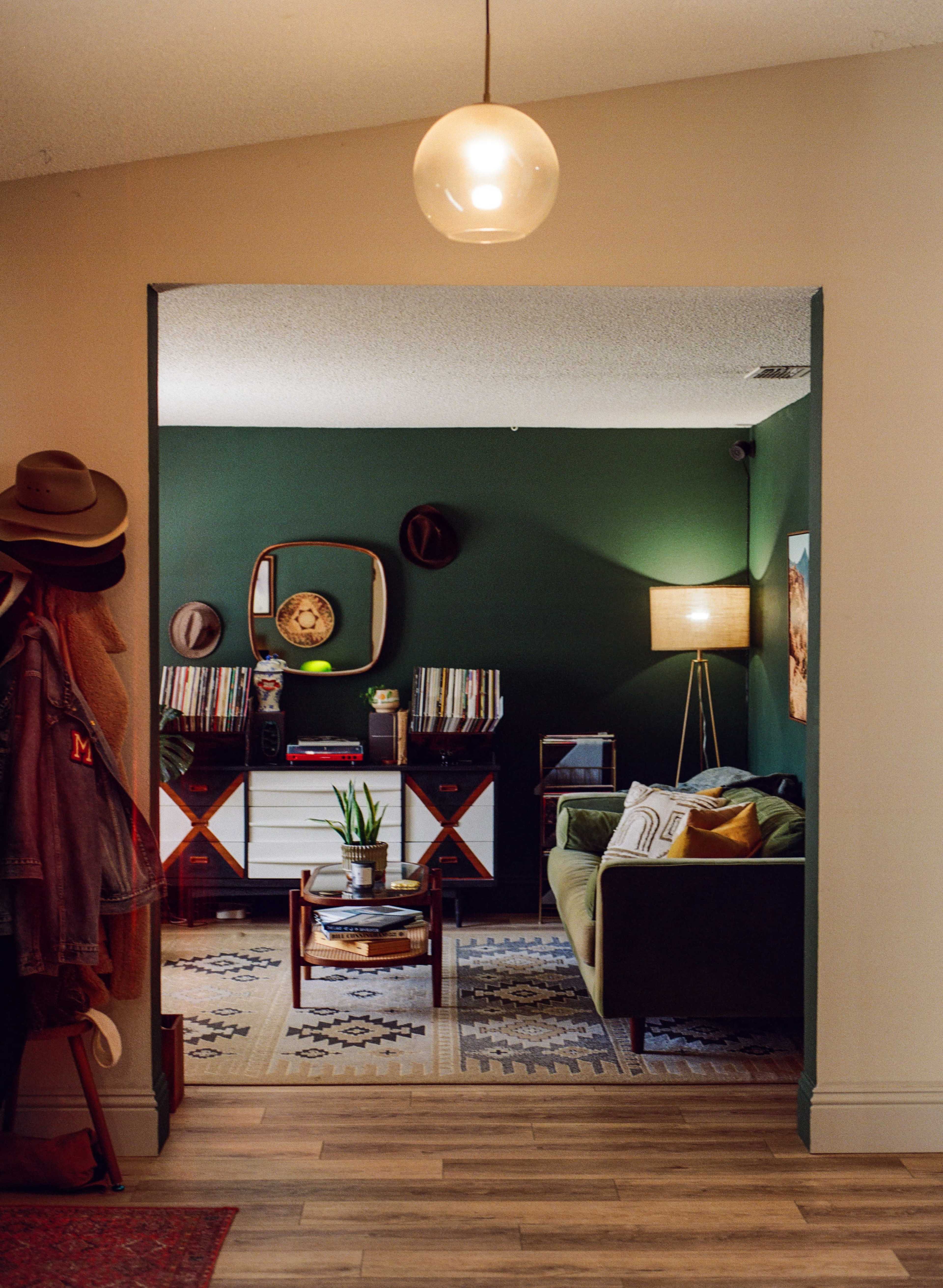 A hallway leads to a cozy living room featuring a green wall, a sofa, a lamp, and shelves filled with vinyl records and hats.