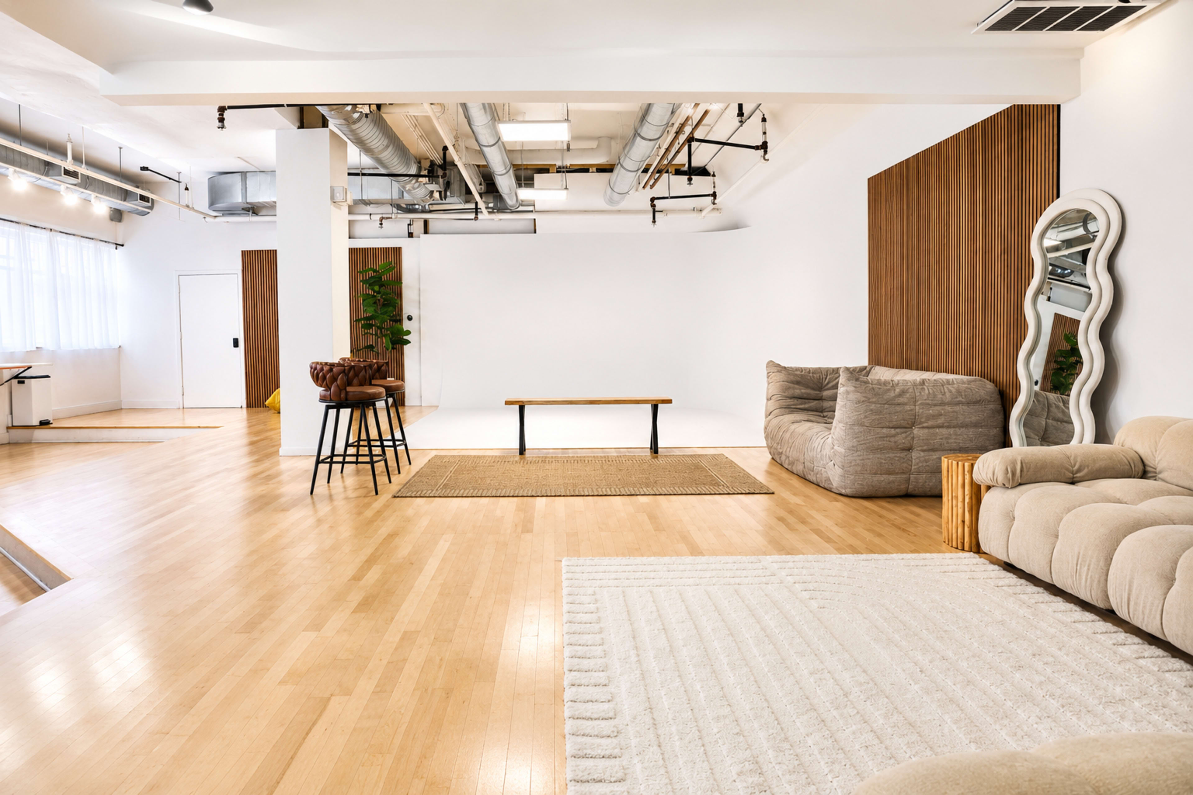 The image shows a spacious, modern interior featuring a light wood floor, a large area rug, a wooden coffee table, a sectional sofa, and a decorative mirror against the wall.