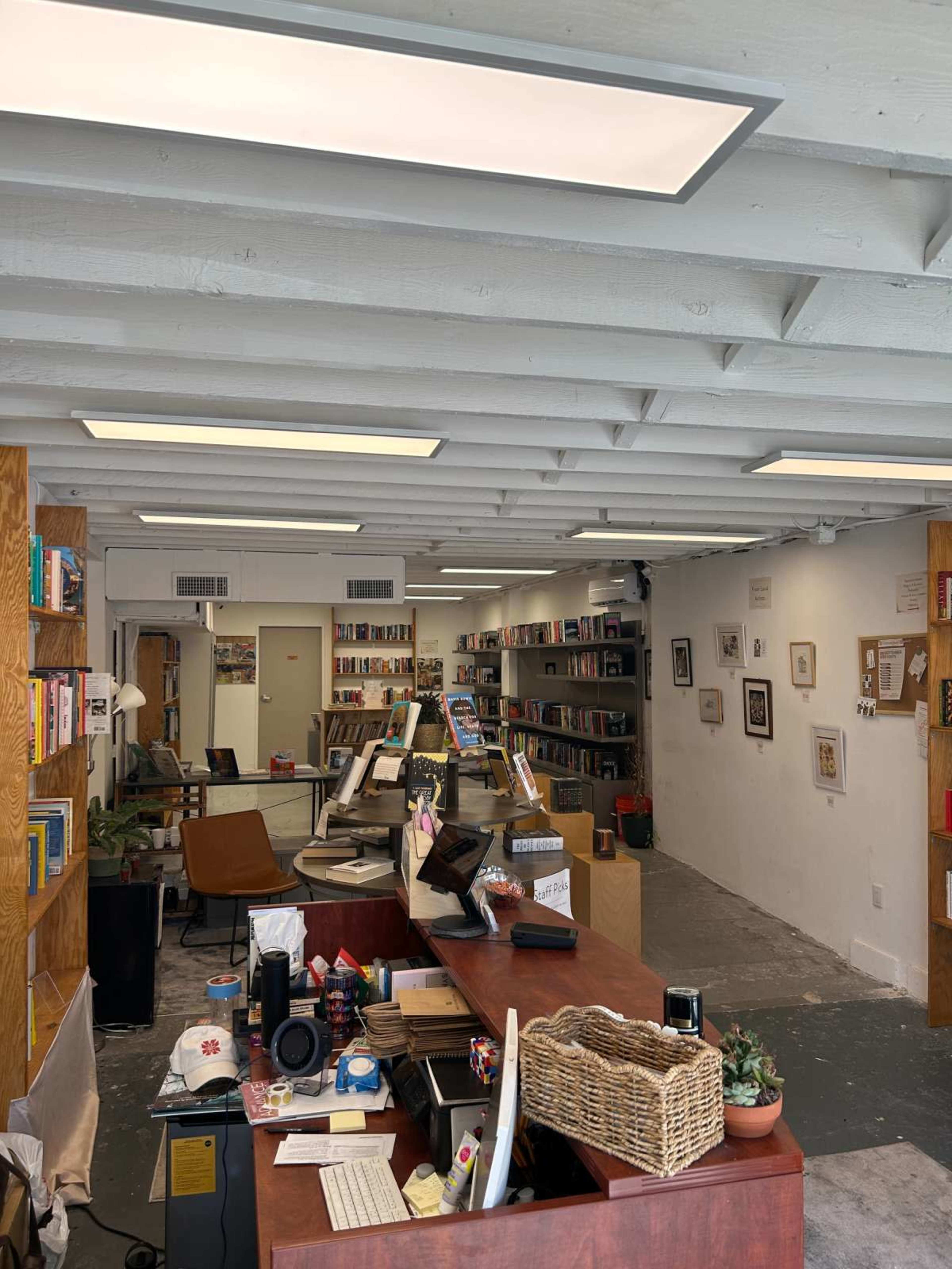 The image shows a spacious bookstore with bookshelves lining the walls, a wooden counter in the foreground, and a cozy seating area.
