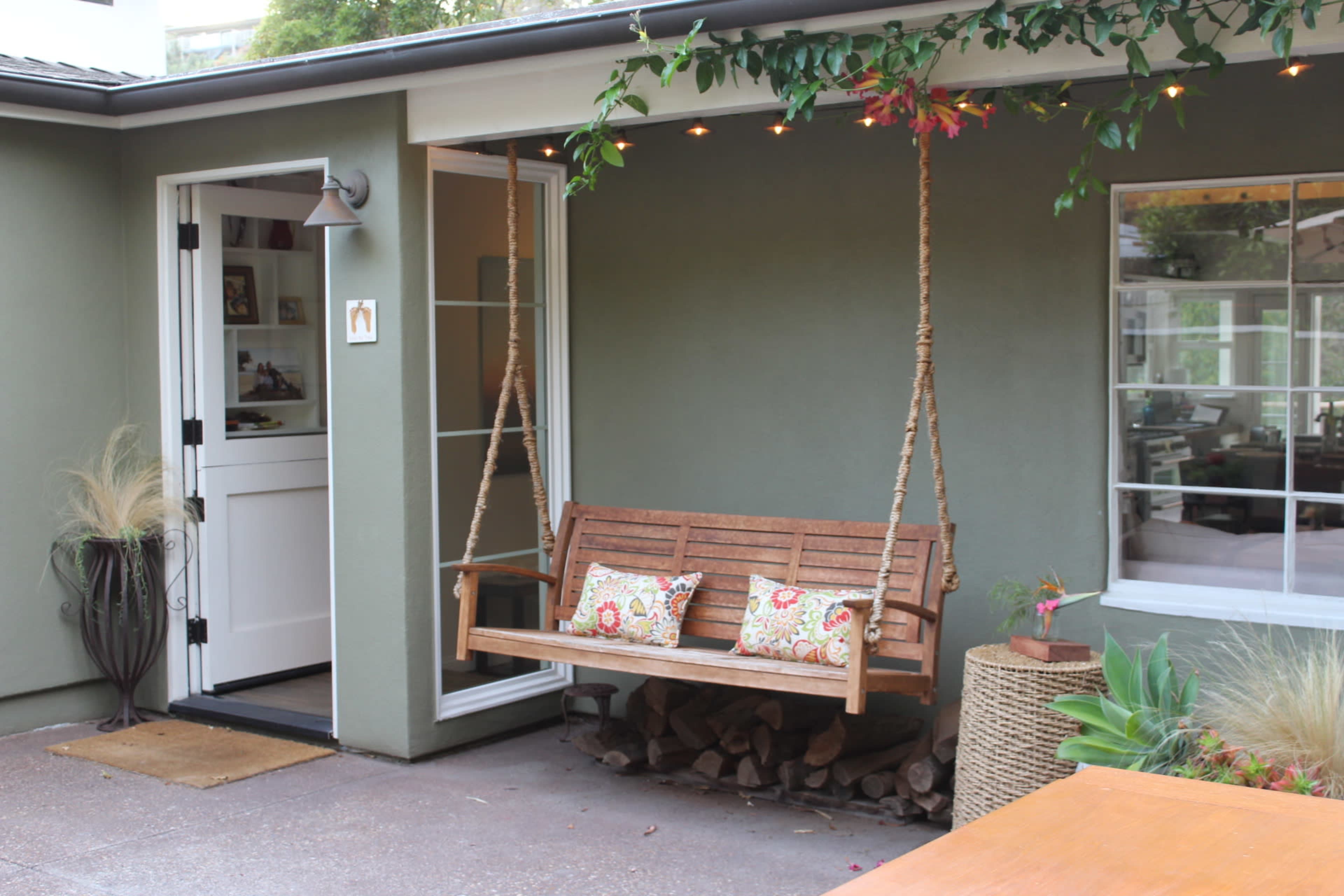 A swing bench with decorative cushions hangs on a porch beside a green wall and large windows.