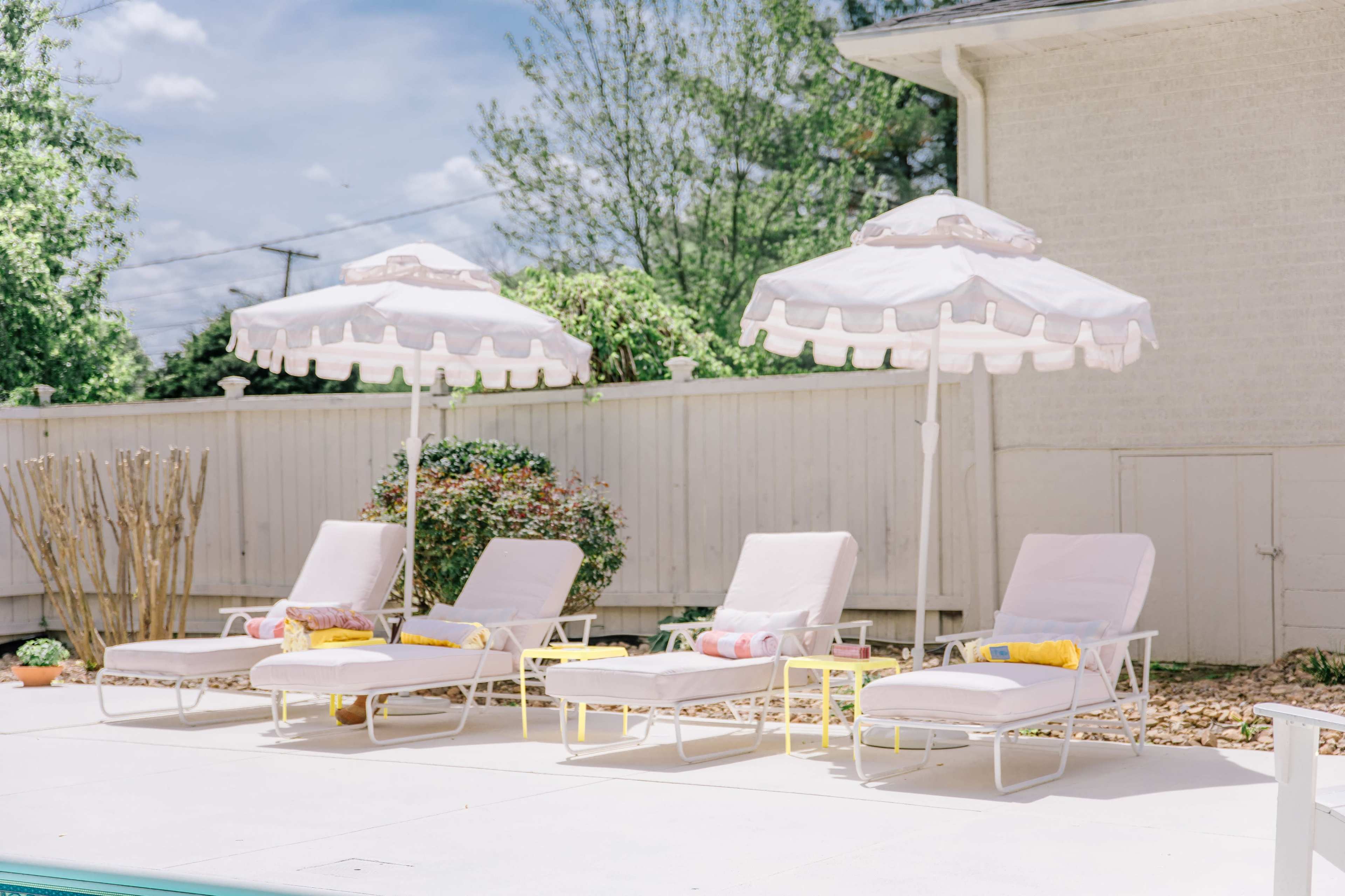 A row of four white lounge chairs with pink cushions and matching umbrellas beside a pool, set against a landscaped backdrop.
