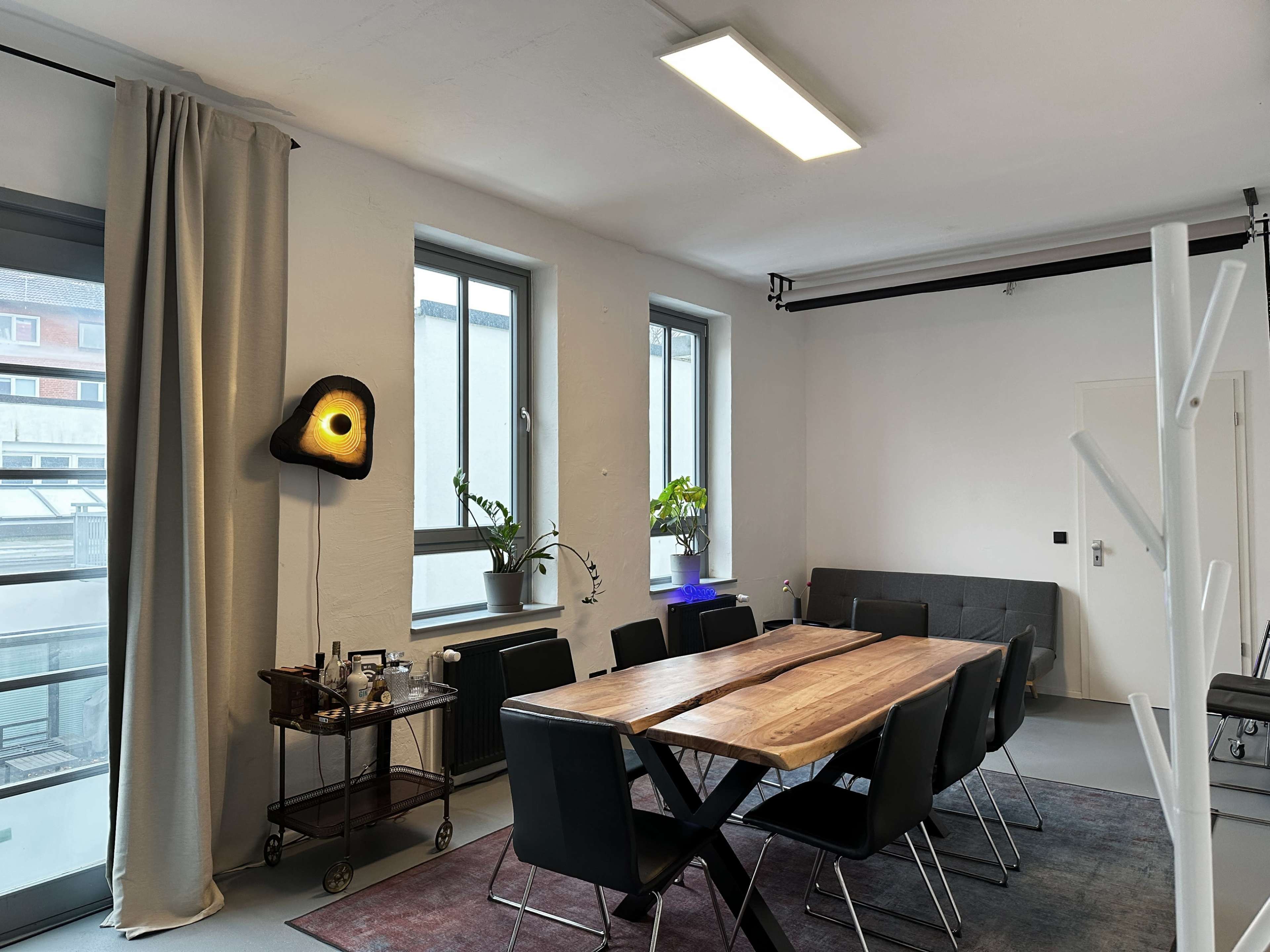 A spacious room features a long wooden dining table surrounded by black chairs, with large windows allowing natural light and a cart displaying beverages in the corner.