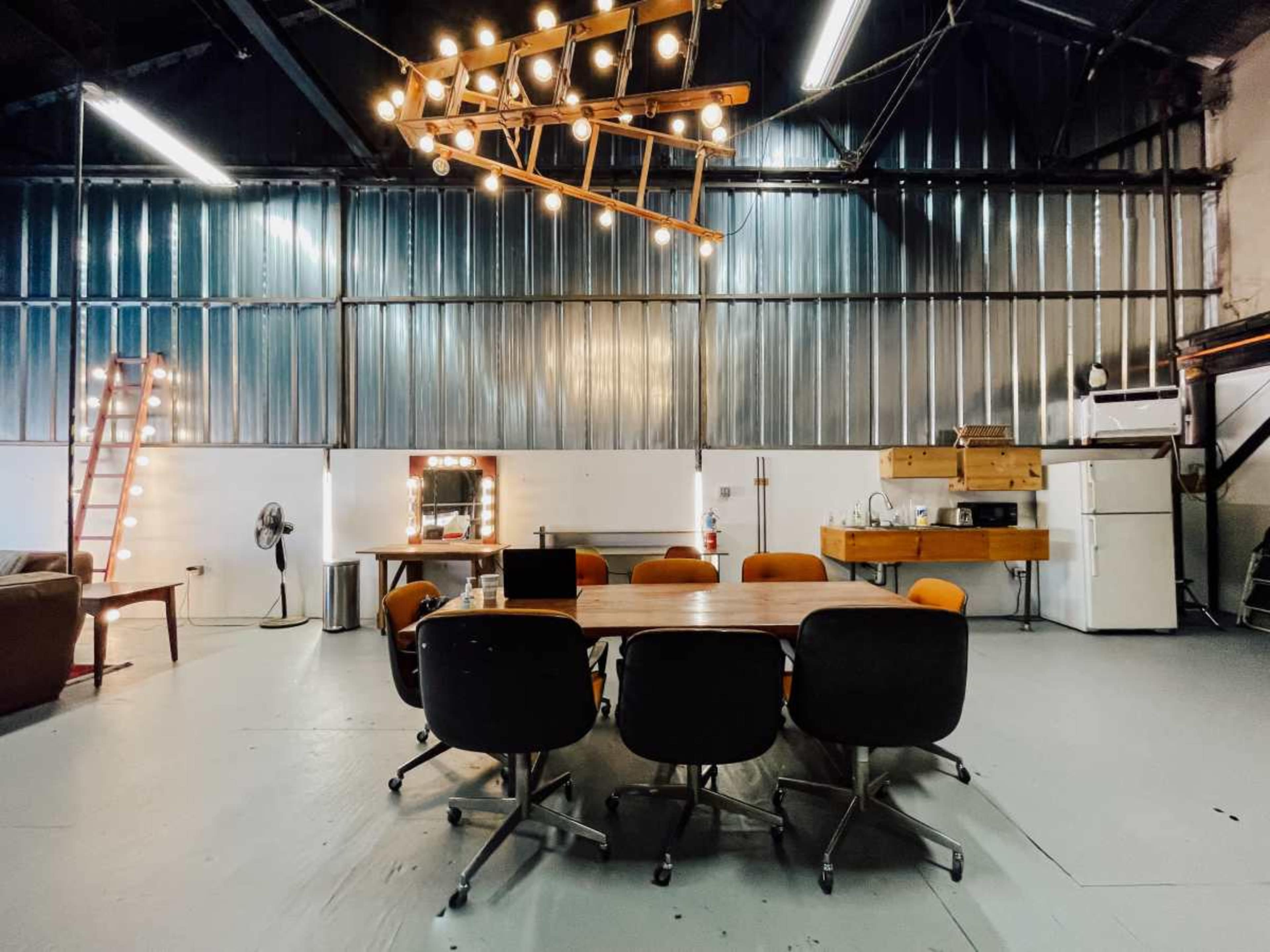 A modern meeting room features a wooden table surrounded by six black chairs, with industrial-style lighting and a kitchenette in the background.