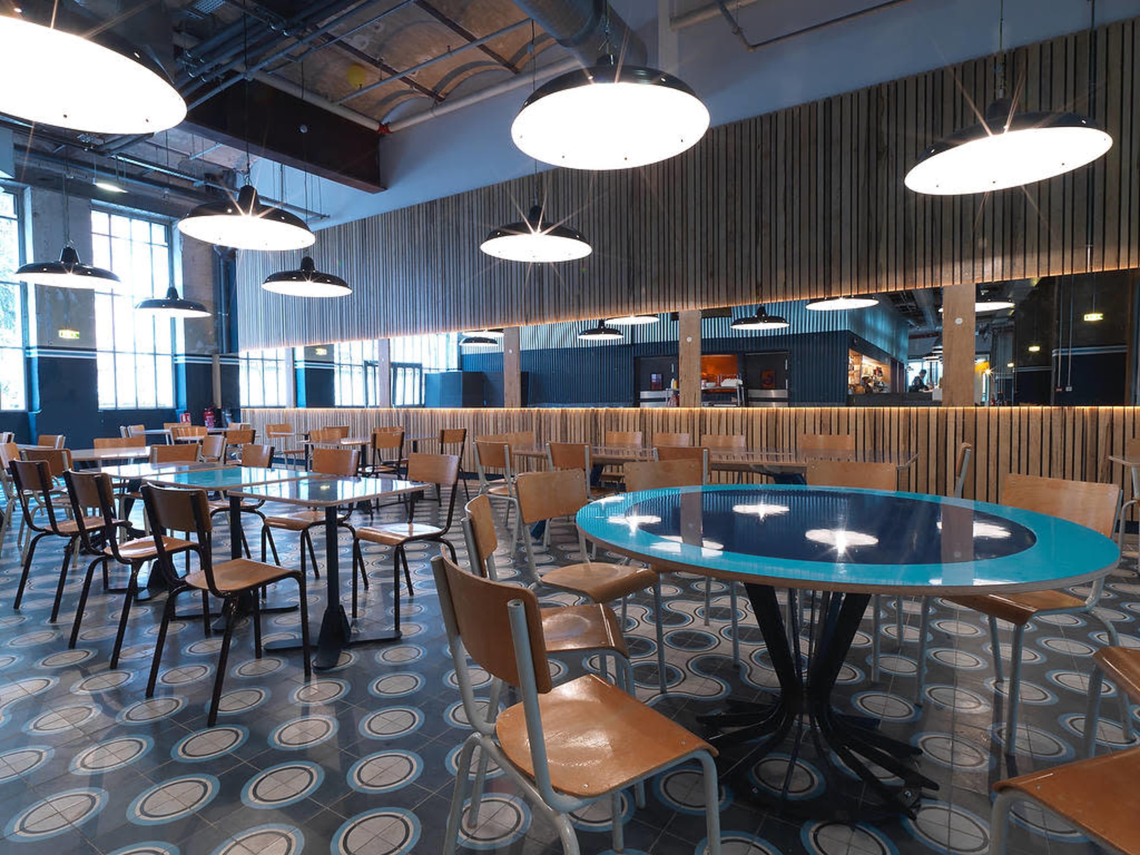 A modern dining area with blue tables, wooden chairs, and overhead lights in a spacious, well-lit room.