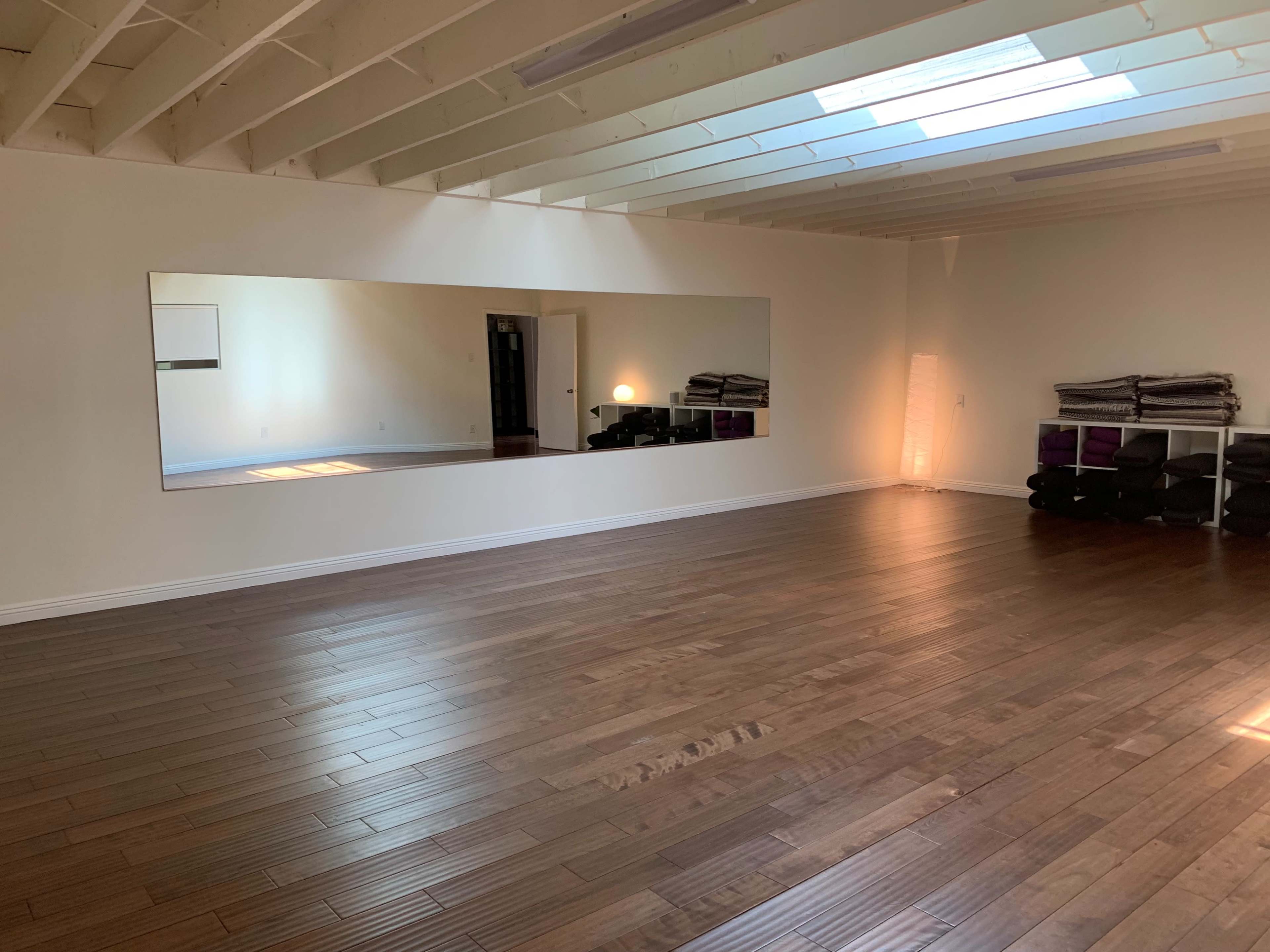 A spacious room with wooden flooring, a large mirror on one wall, and neatly stacked yoga mats in the corner.