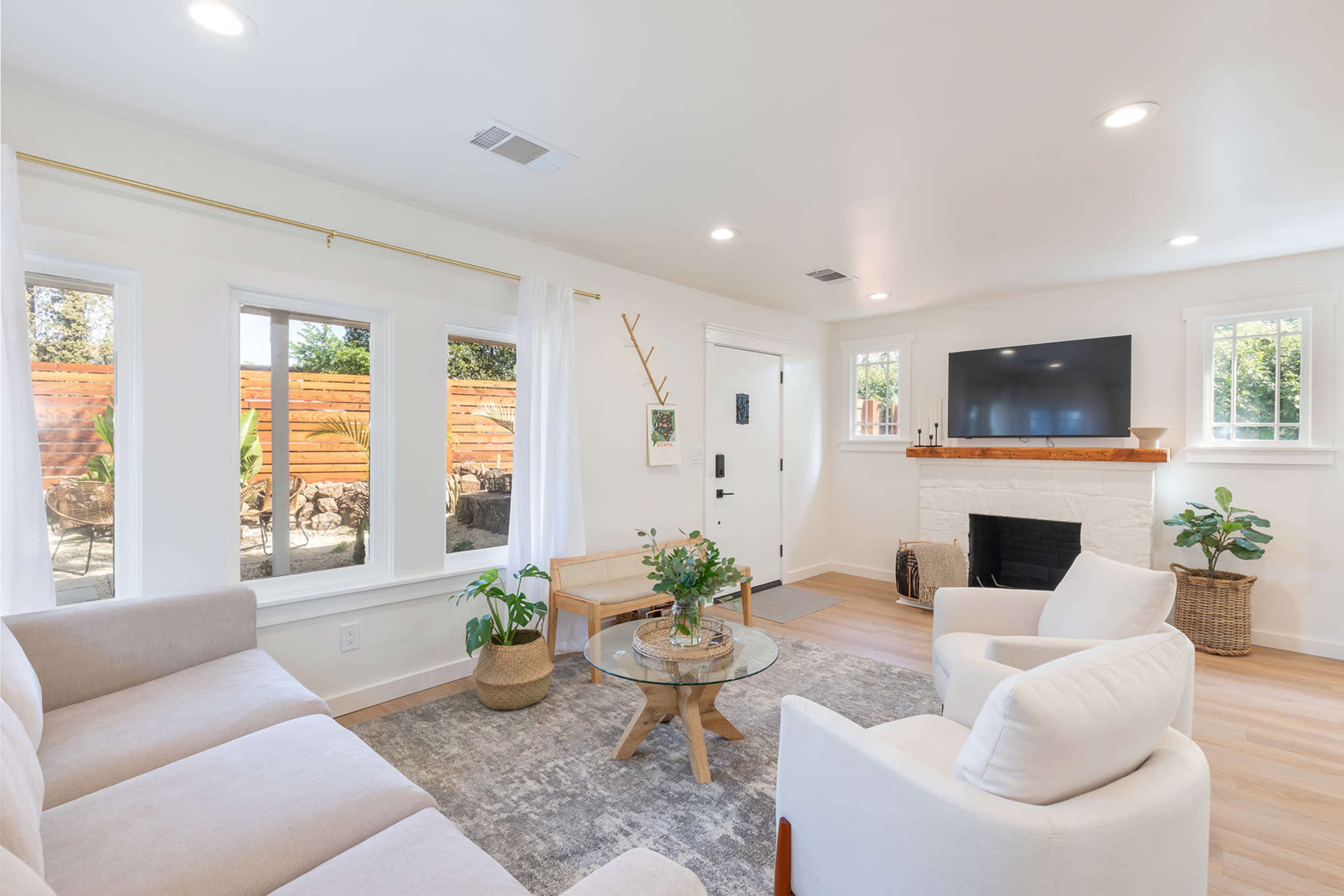 The living room features a light-colored sofa set, a glass coffee table, and a television mounted above a white stone fireplace, with large windows allowing natural light to enter.