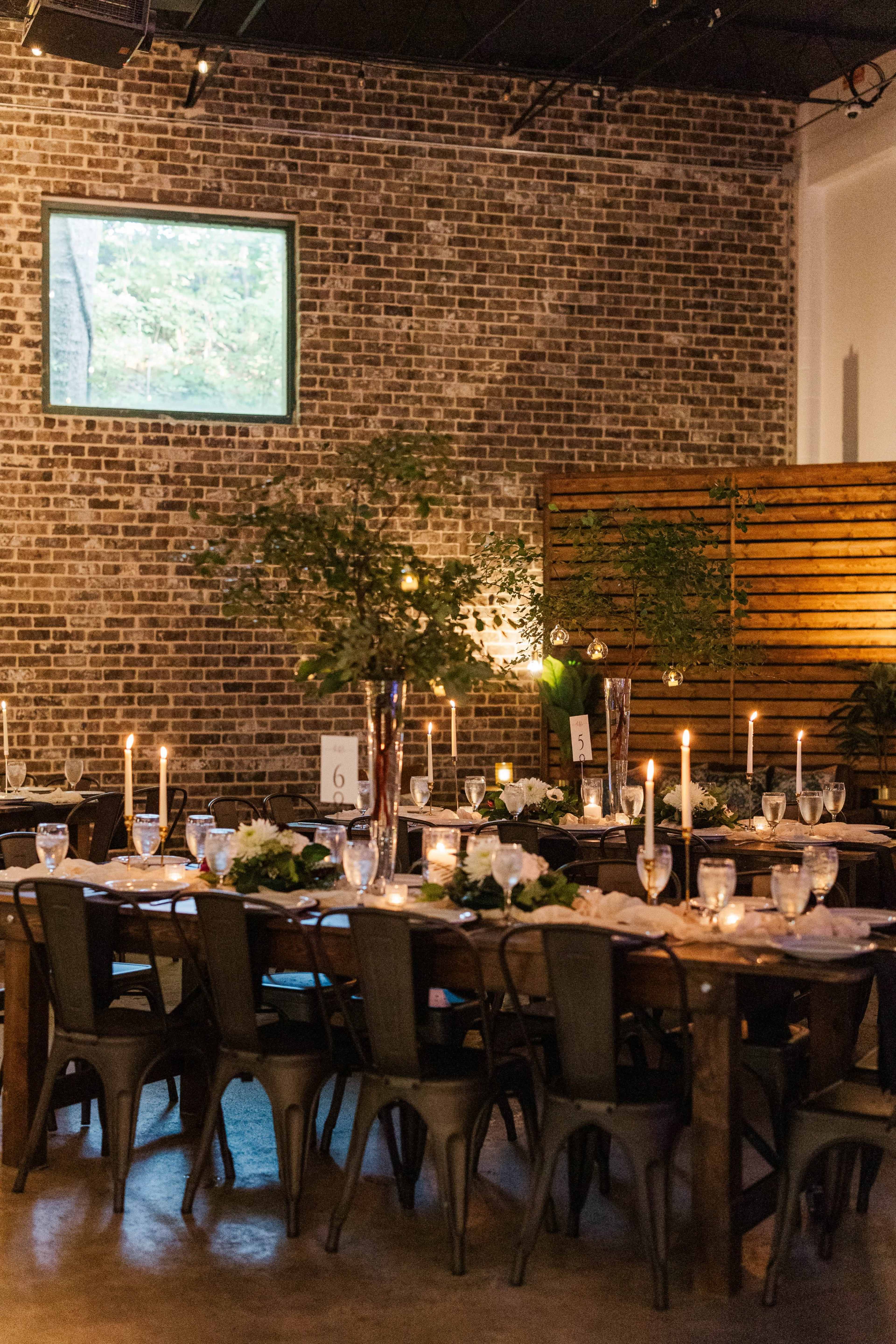 A rustic dining area features wooden tables set with elegant floral centerpieces, candlelight, and a backdrop of exposed brick walls.