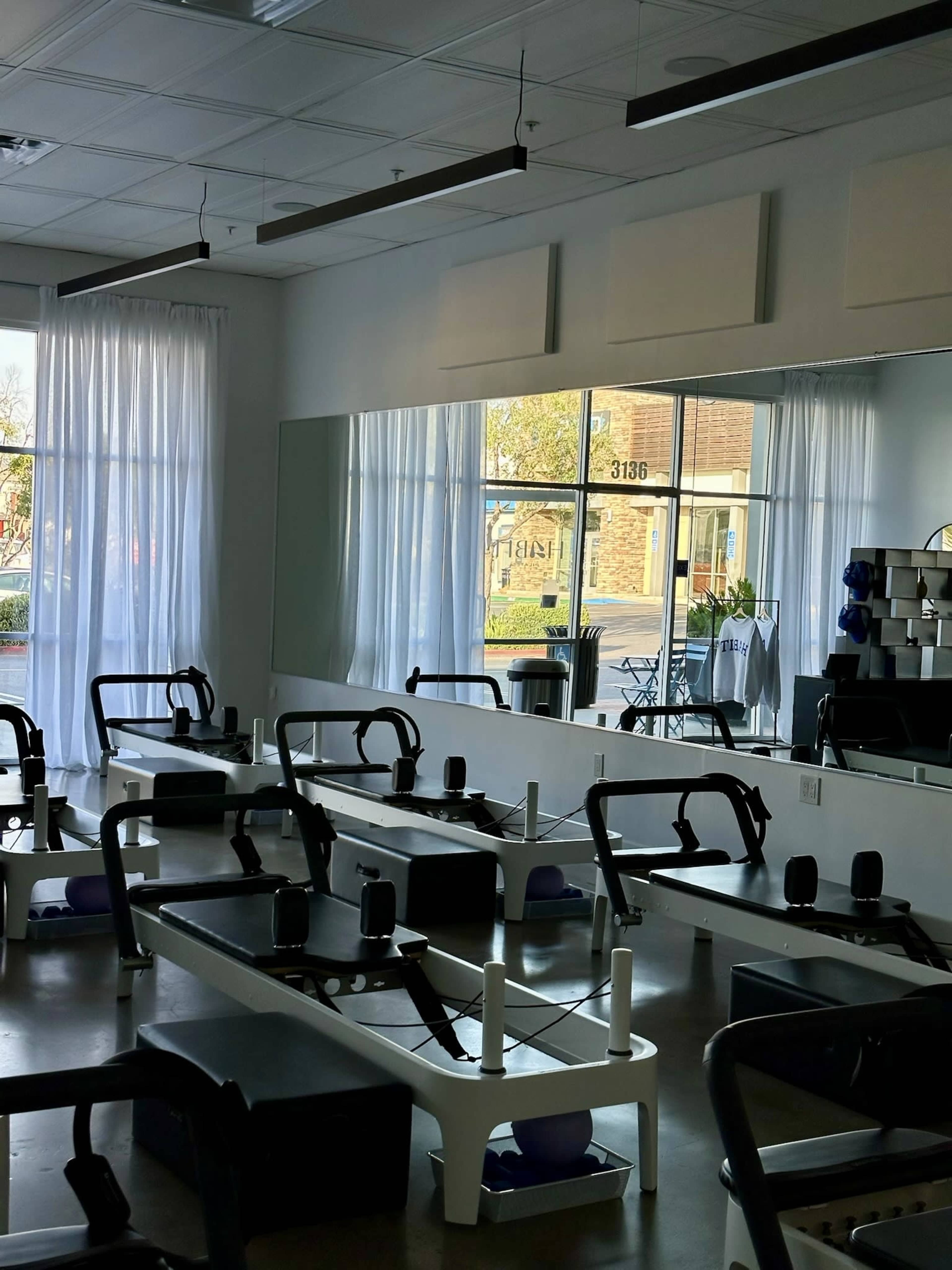 The image shows a modern fitness studio with reformer machines arranged neatly in rows, large windows letting in natural light, and mirrors reflecting the interior.