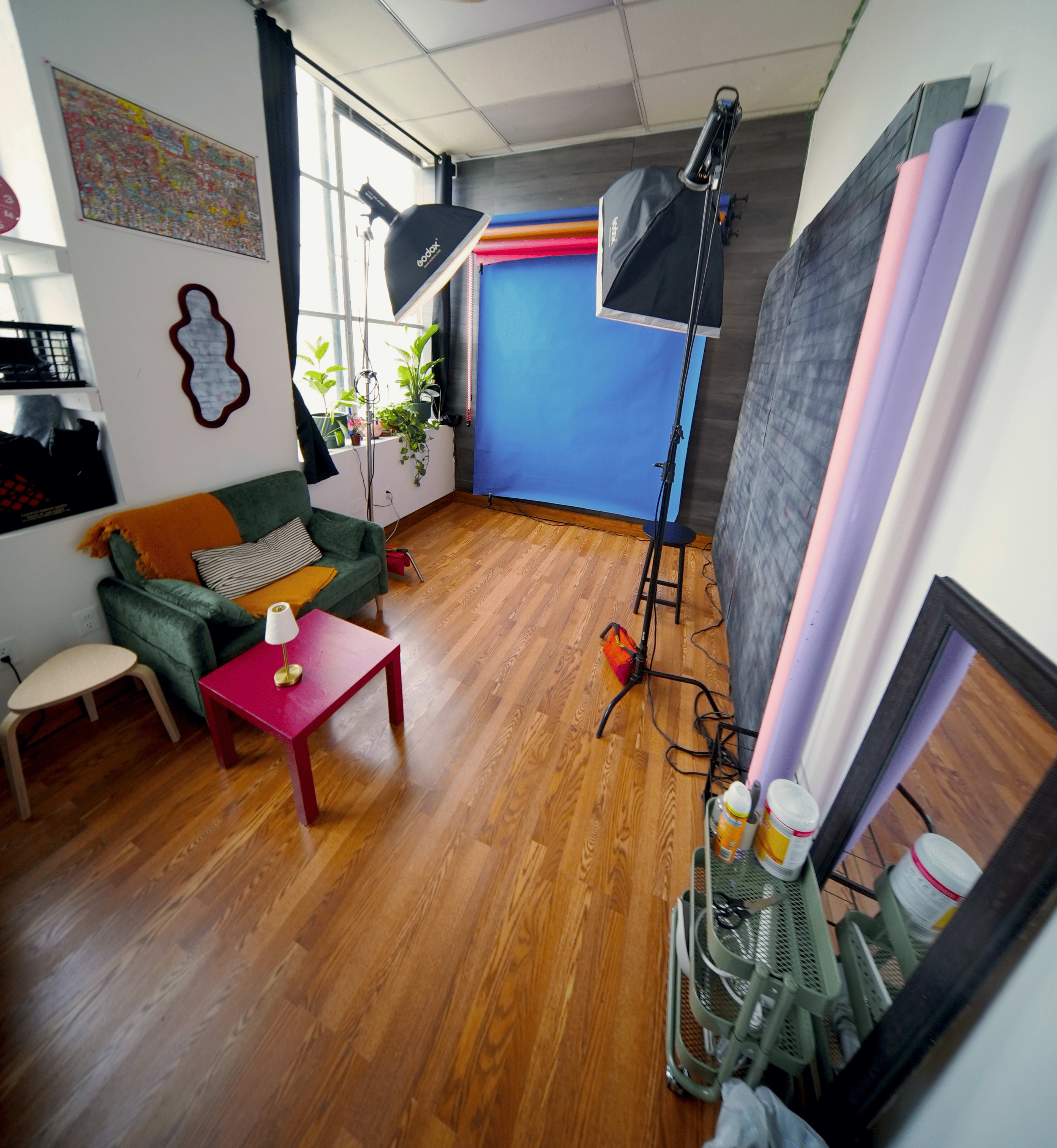 The image shows a bright, spacious studio setup with a green couch, a pink table, photography lights, and a colorful backdrop against a wooden floor.