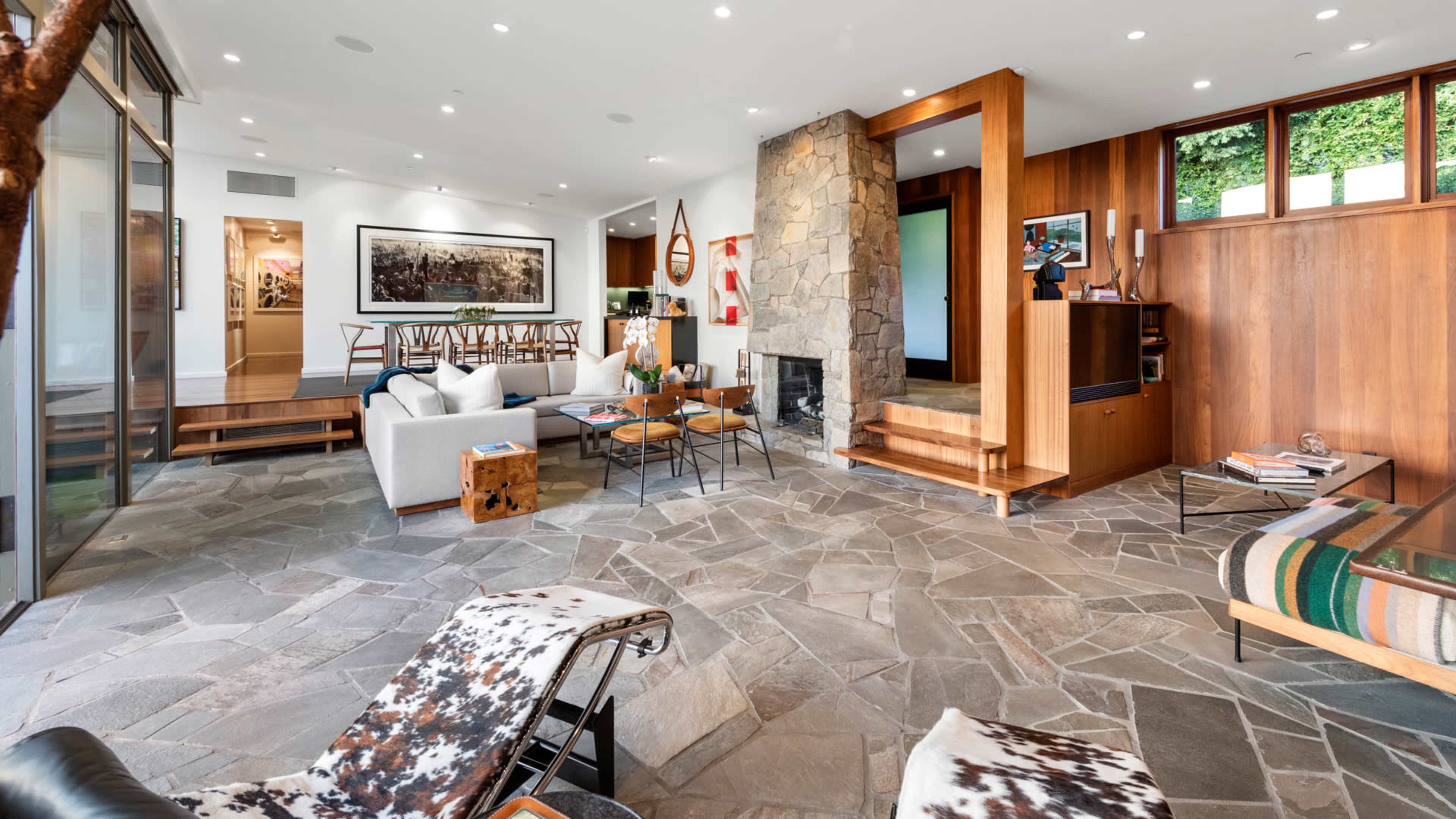 The image depicts a modern living room with a stone fireplace, large windows, and a combination of wooden and stone elements in its design.