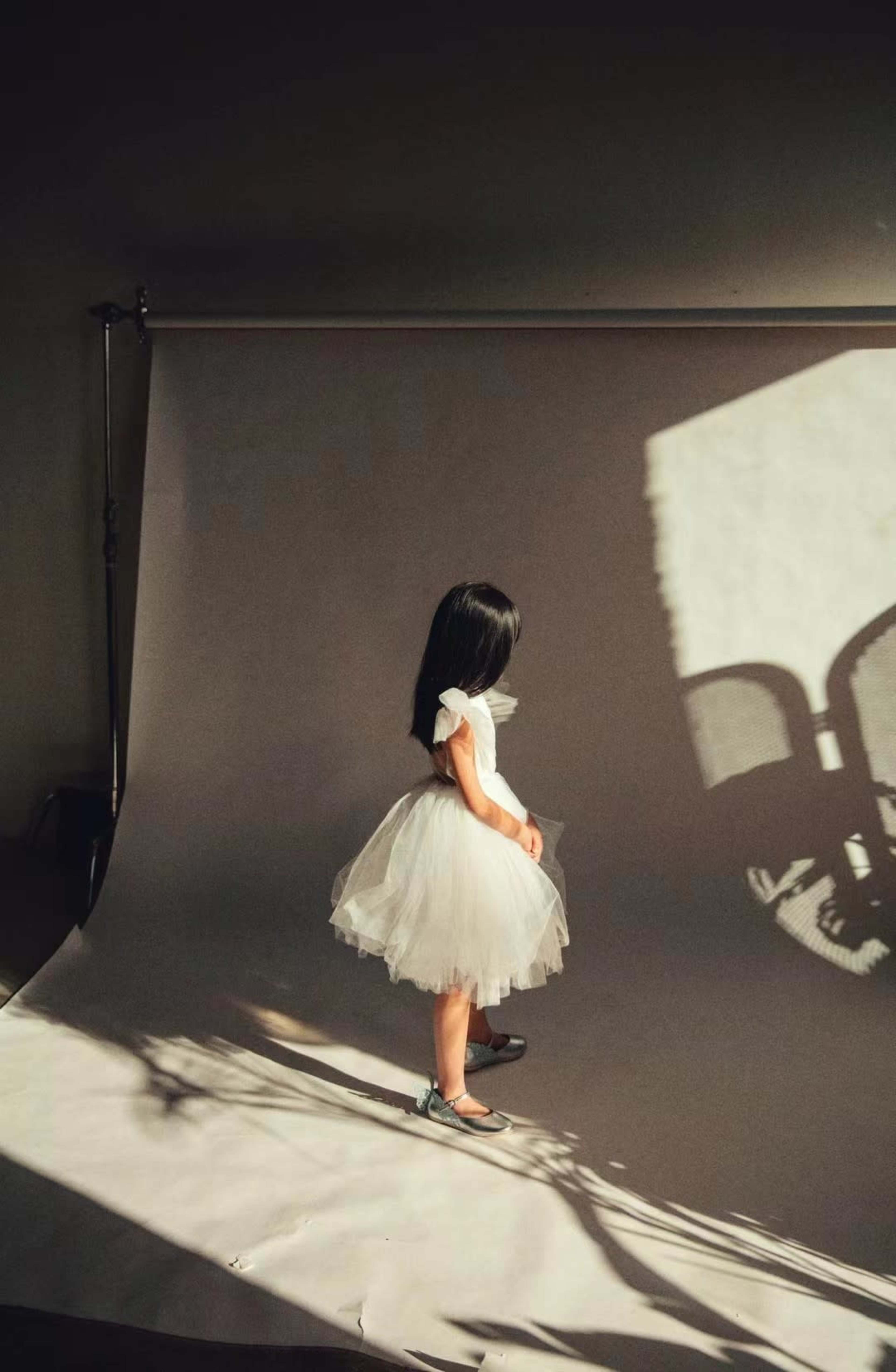 A young girl in a white dress stands on a gray backdrop, with shadows cast across the background and floor.