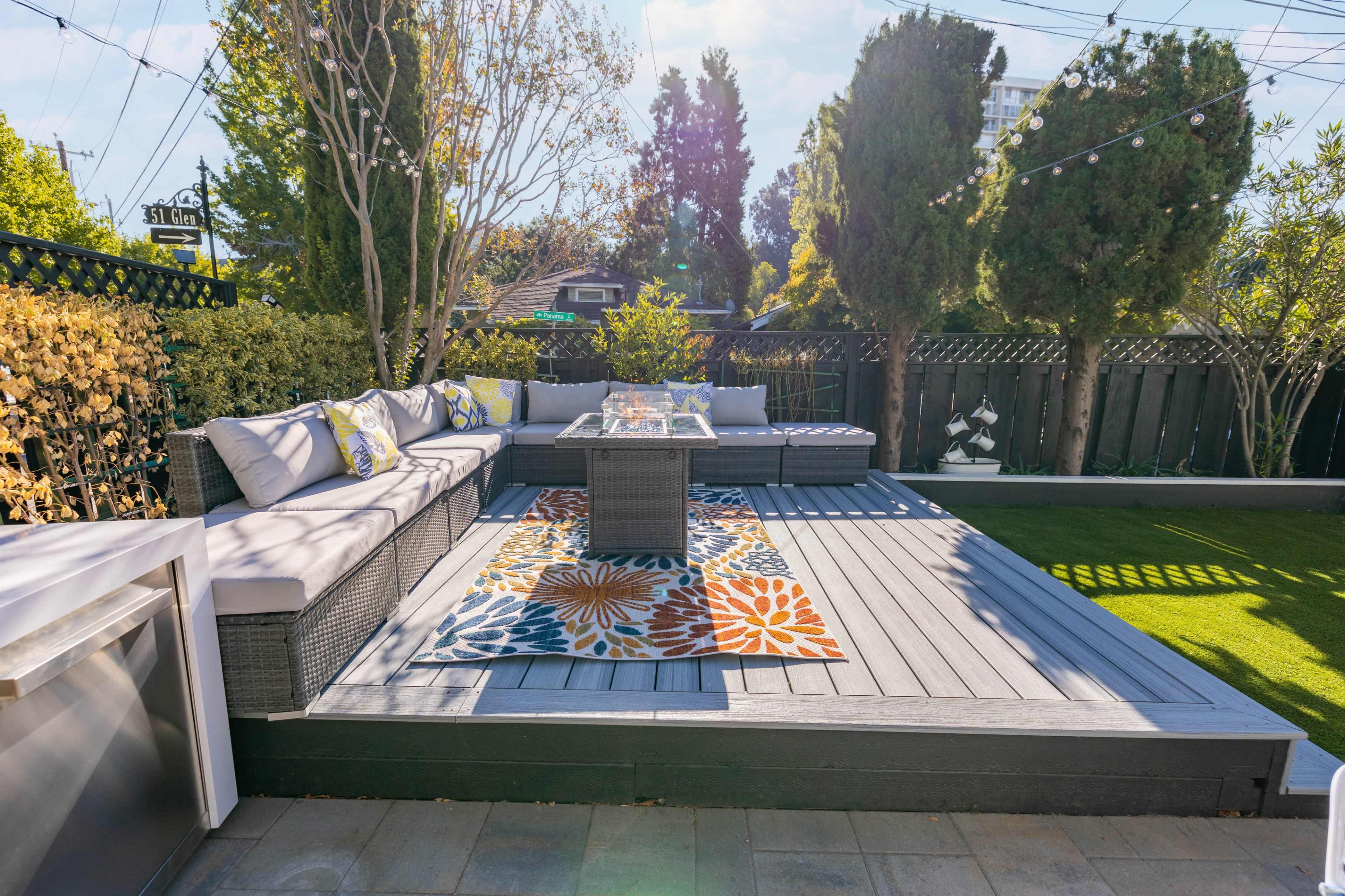 A patio area featuring a gray deck with a seating arrangement, a central table, and a colorful rug surrounded by greenery.
