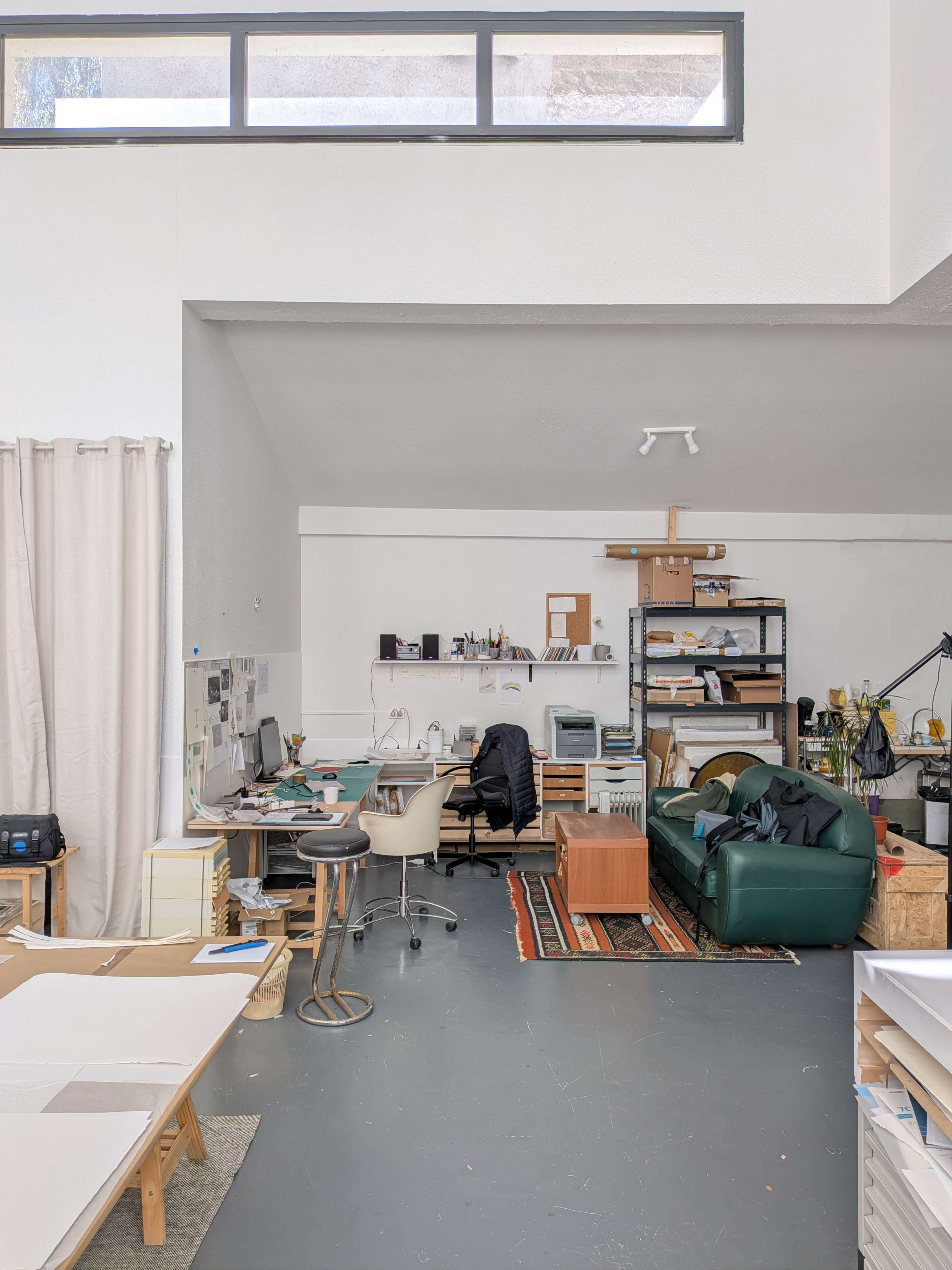 The image shows a spacious studio with a desk and computer, a green couch, a bookshelf filled with supplies, and large windows providing natural light.