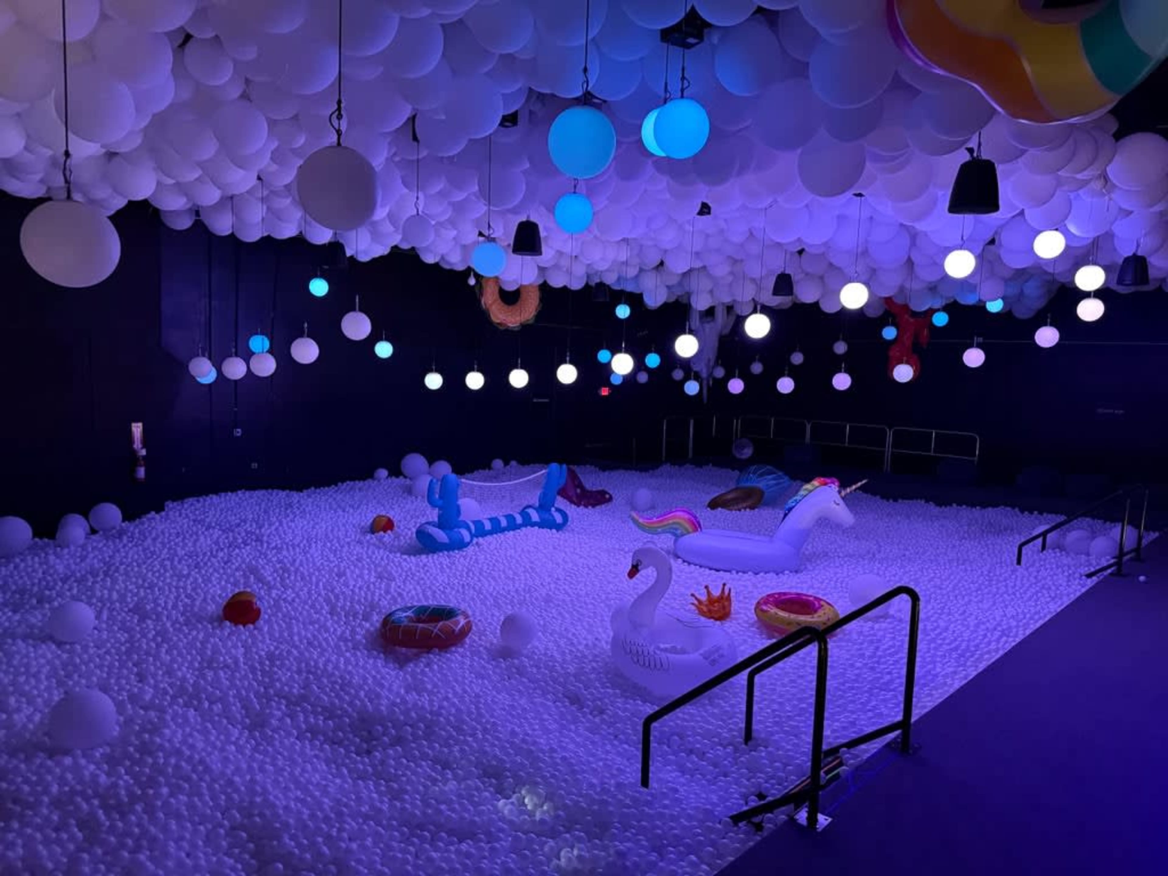 The room is filled with plastic balls and decorated with inflated floating toys under a canopy of balloons and colorful lights.