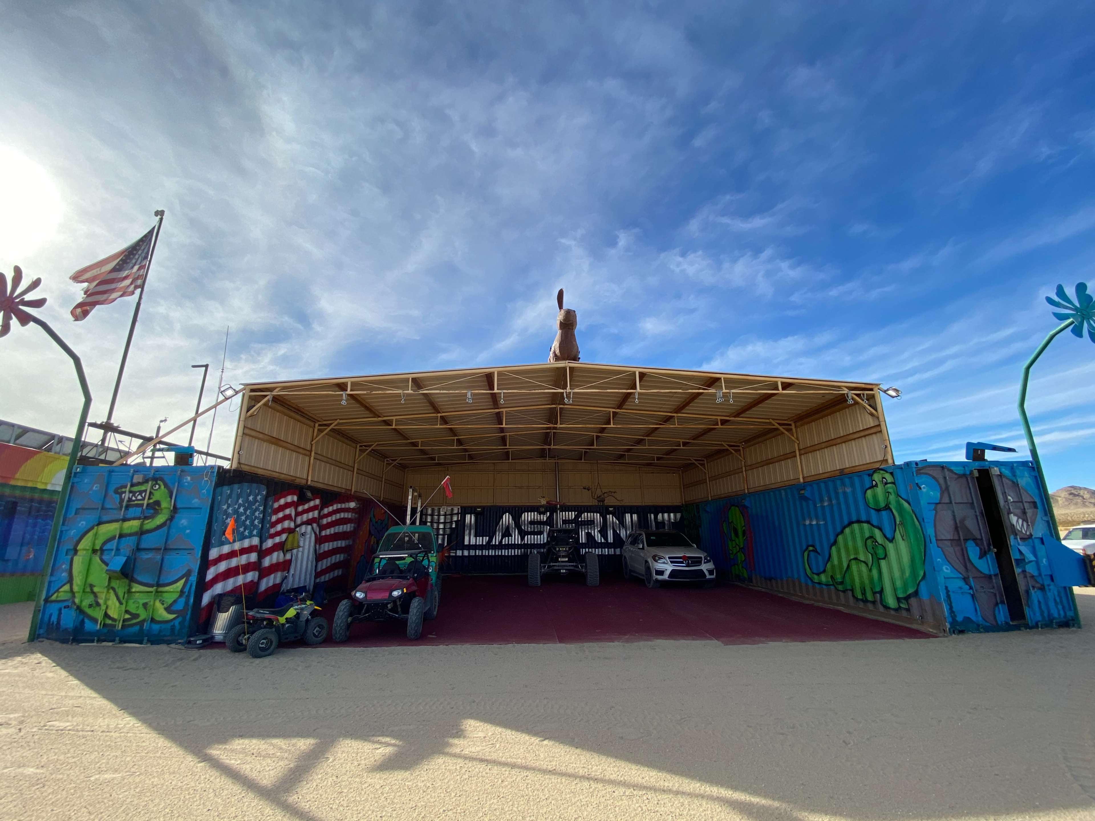 The image shows a colorful building with murals, featuring a large dinosaur and American flags, housing vehicles and a sculpture of a rabbit on the roof.