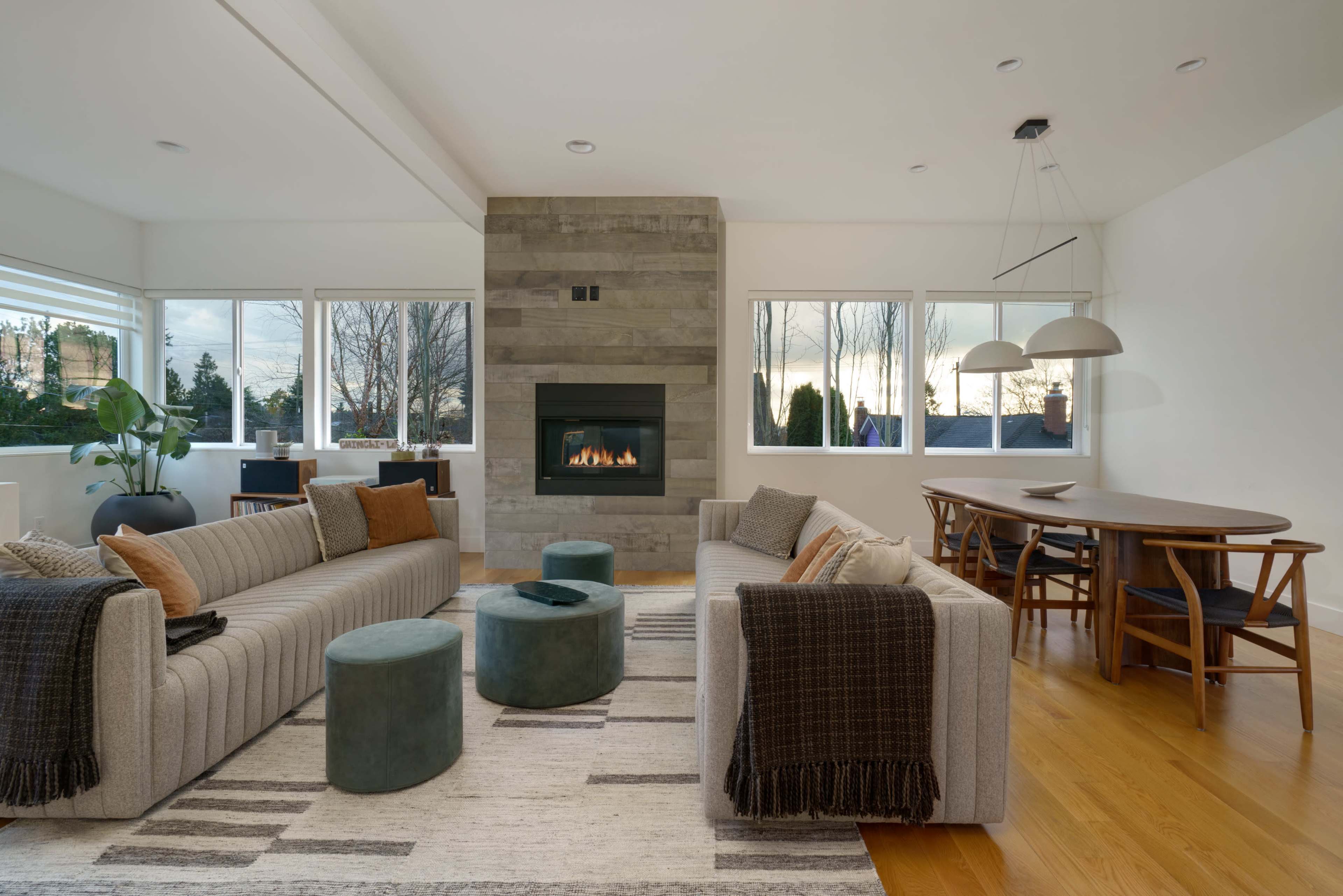 A modern living room features two gray sofas, a circular dining table, a stone fireplace, and large windows with a view of trees outside.