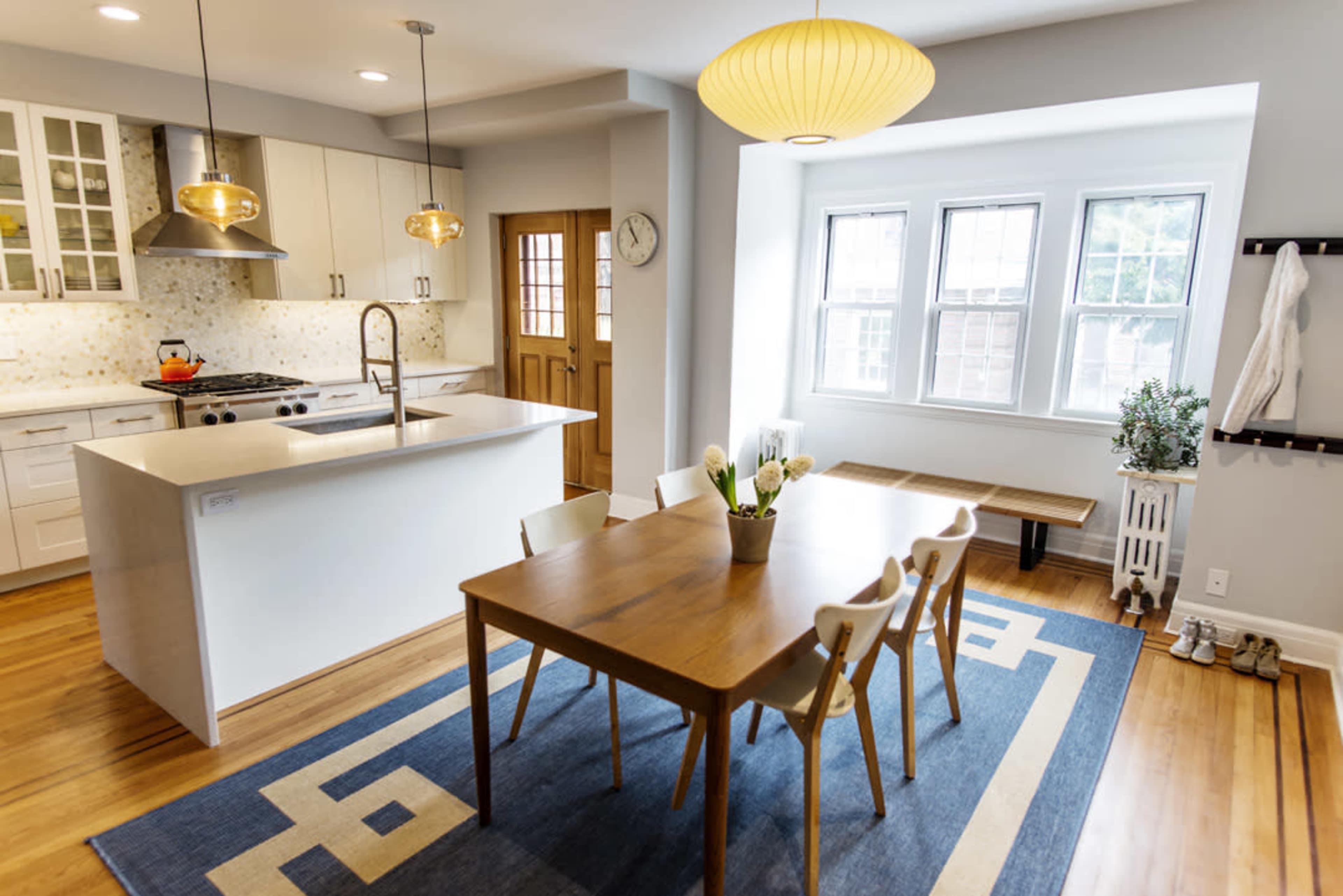 A modern kitchen and dining area features a wooden table set with chairs, pendant lighting, and large windows allowing natural light to fill the space.