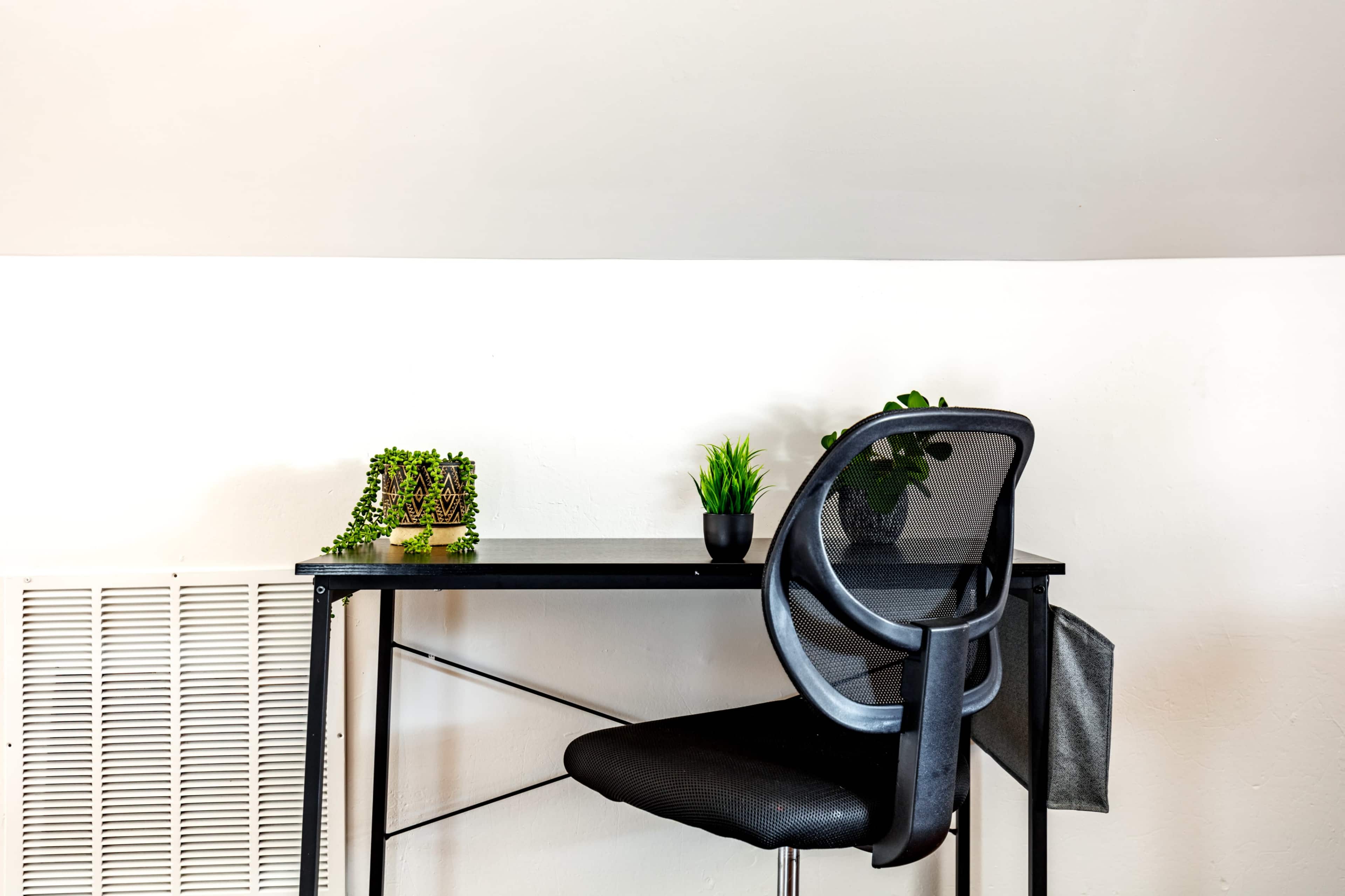 A black desk with a mesh chair and two small potted plants is positioned against a white wall.