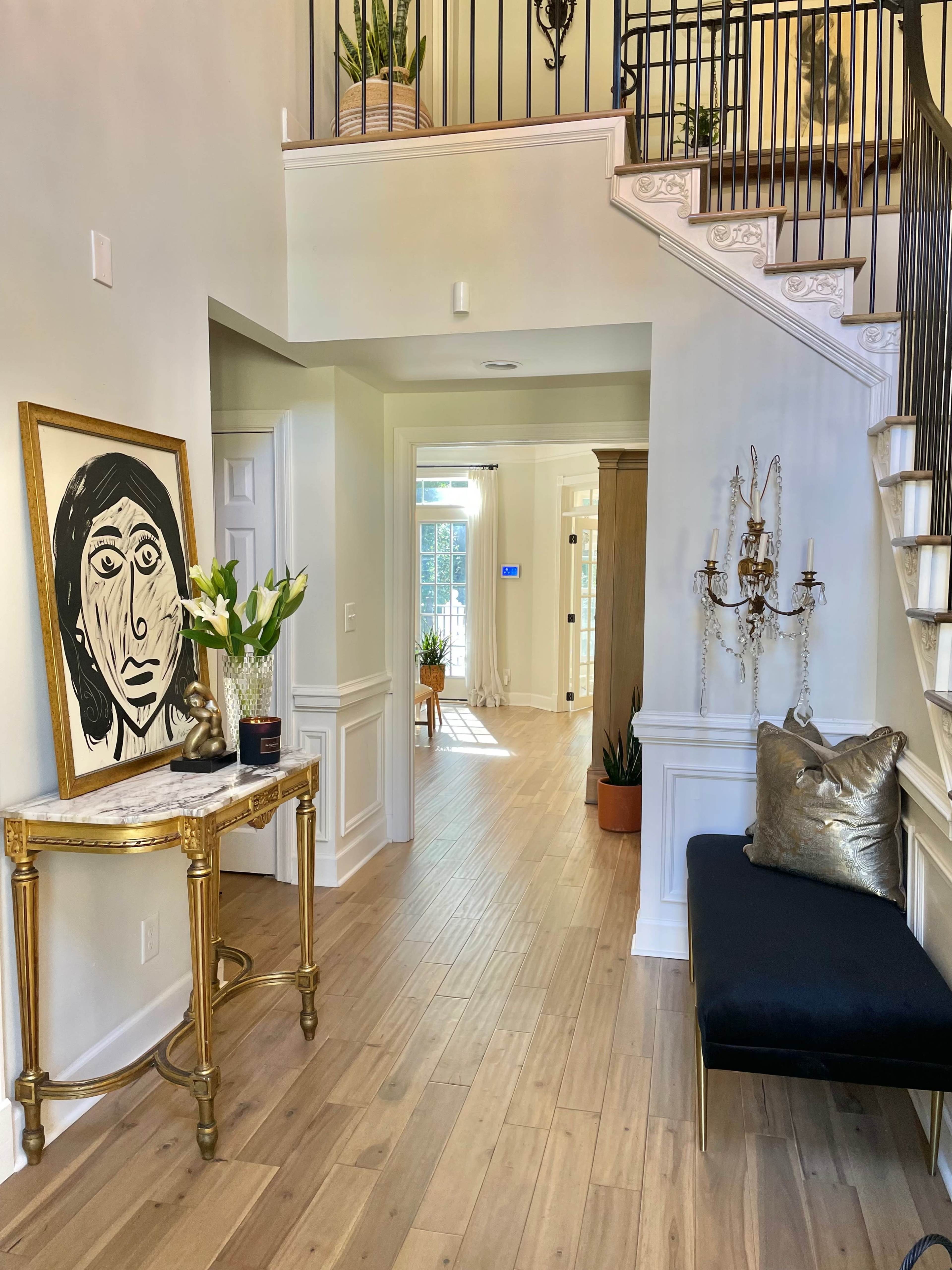 A foyer with a staircase, a decorative table holding a large framed artwork, and potted plants along the walls.