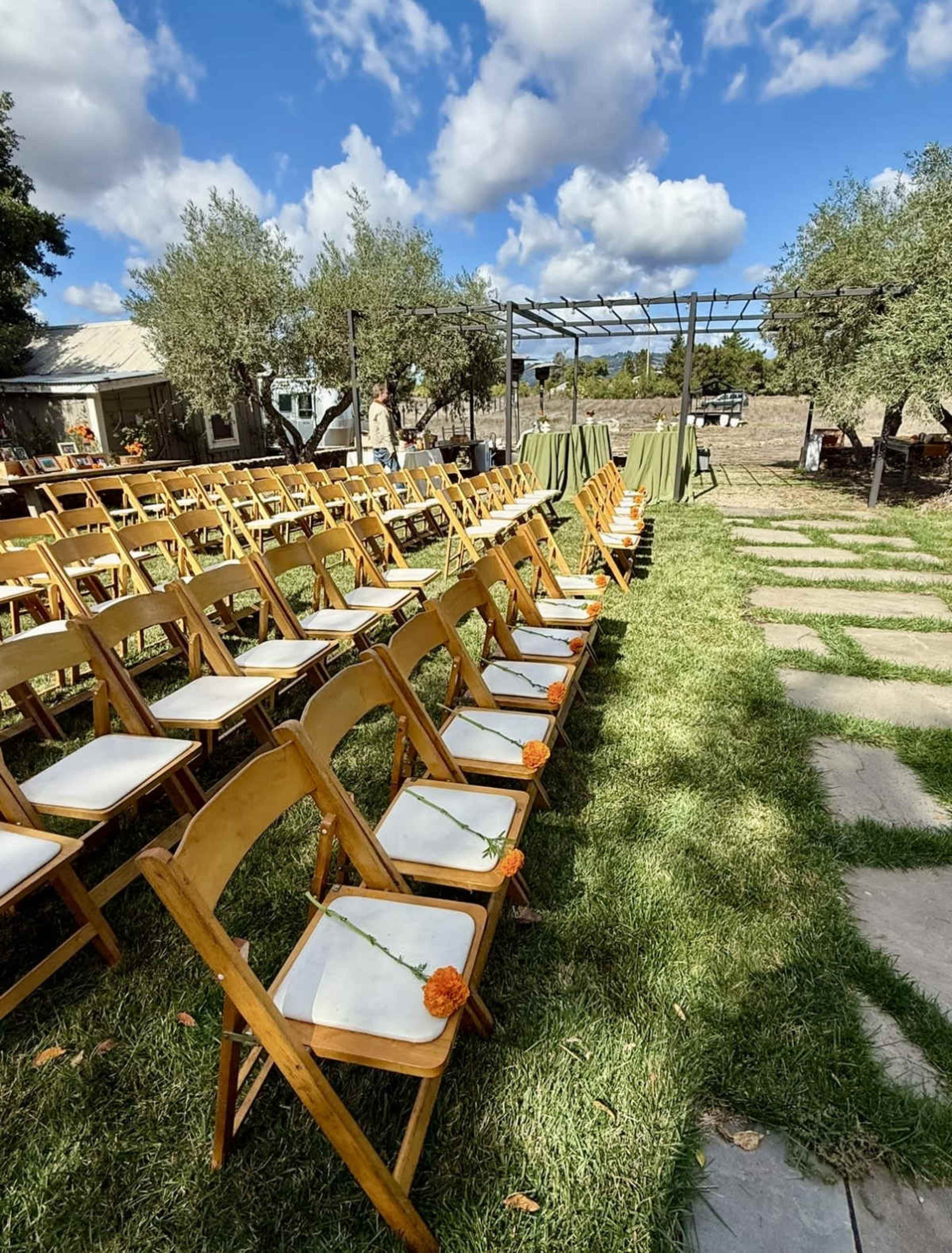 The image shows a row of wooden chairs arranged for an outdoor event, with a path of stone tiles leading toward a structure in the background, under a partly cloudy sky.