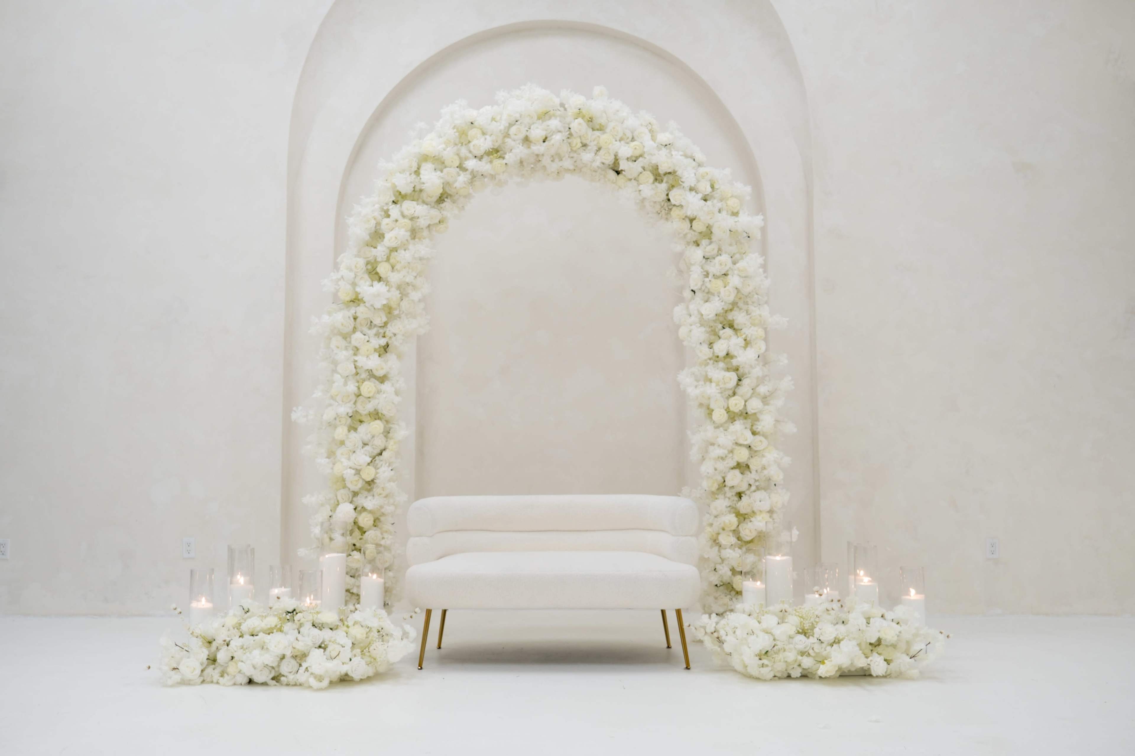 A cream-colored sofa sits beneath a floral arch adorned with white flowers, surrounded by arrangements of blooms and candles on a light-colored floor.