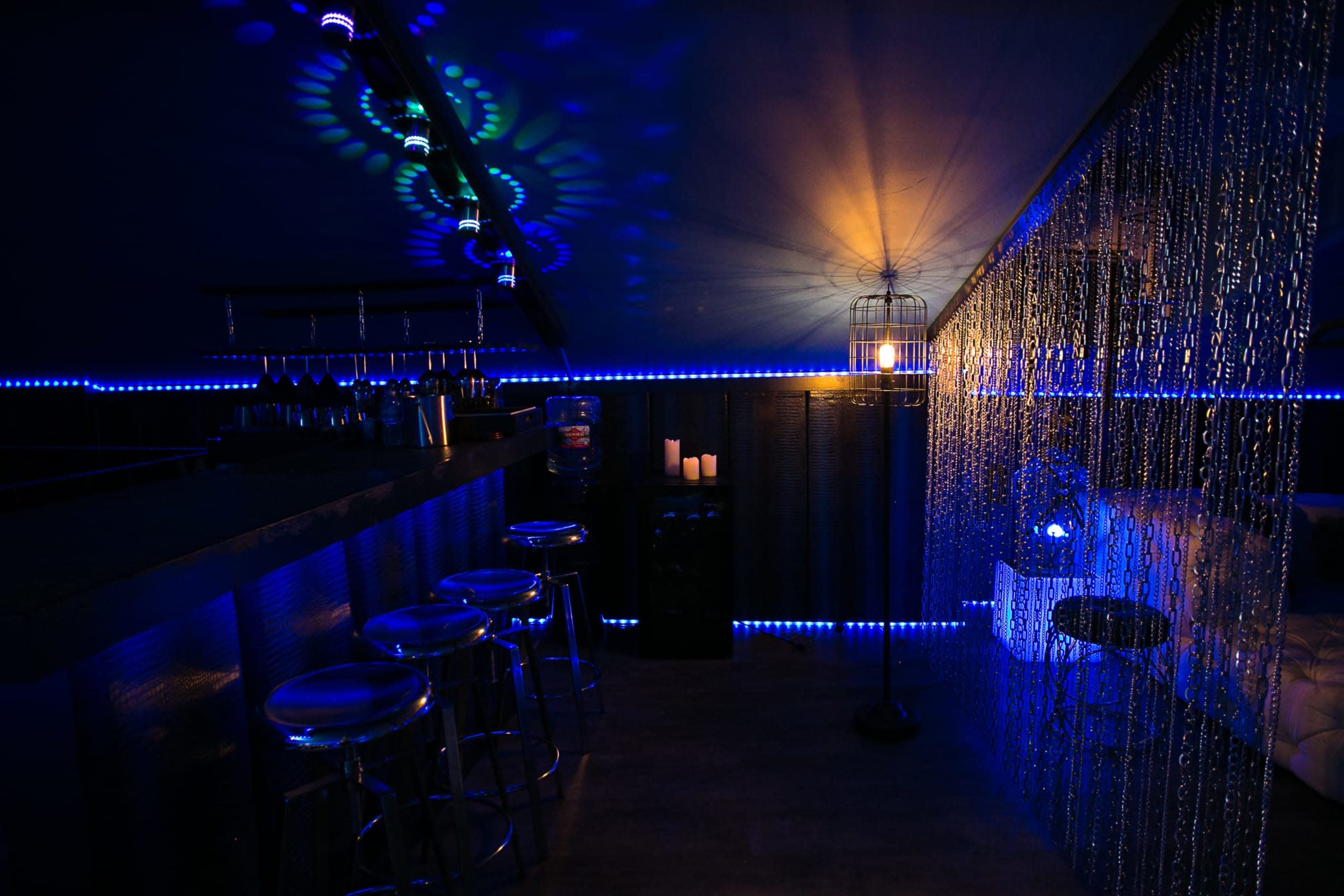 A dimly lit bar area with blue lighting, a counter with stools, and decorative chain curtains beside a cage-like lantern.