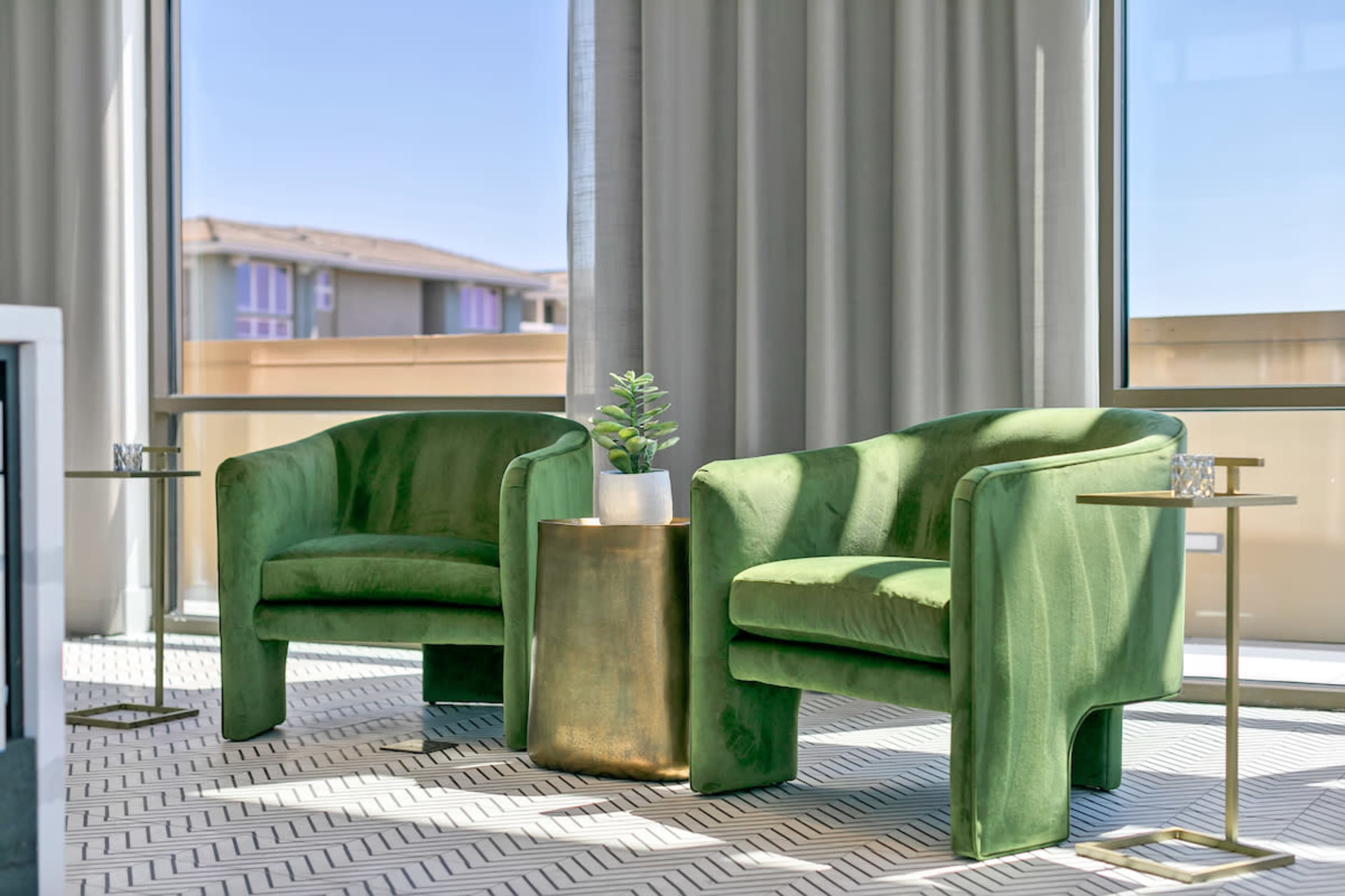 Two green velvet chairs flanking a small gold side table, with a potted plant, set against large windows and sheer curtains.