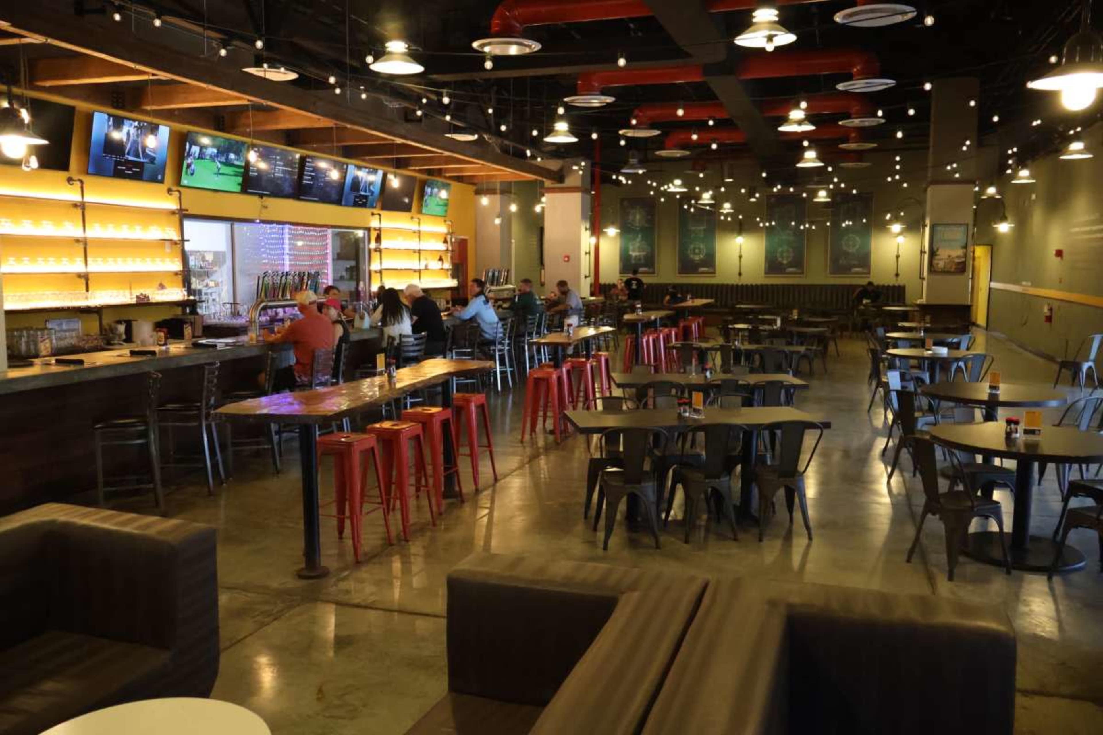 The image shows a spacious, modern restaurant with a bar area, seating arrangements of tables and chairs, and dim lighting complemented by hanging lights.