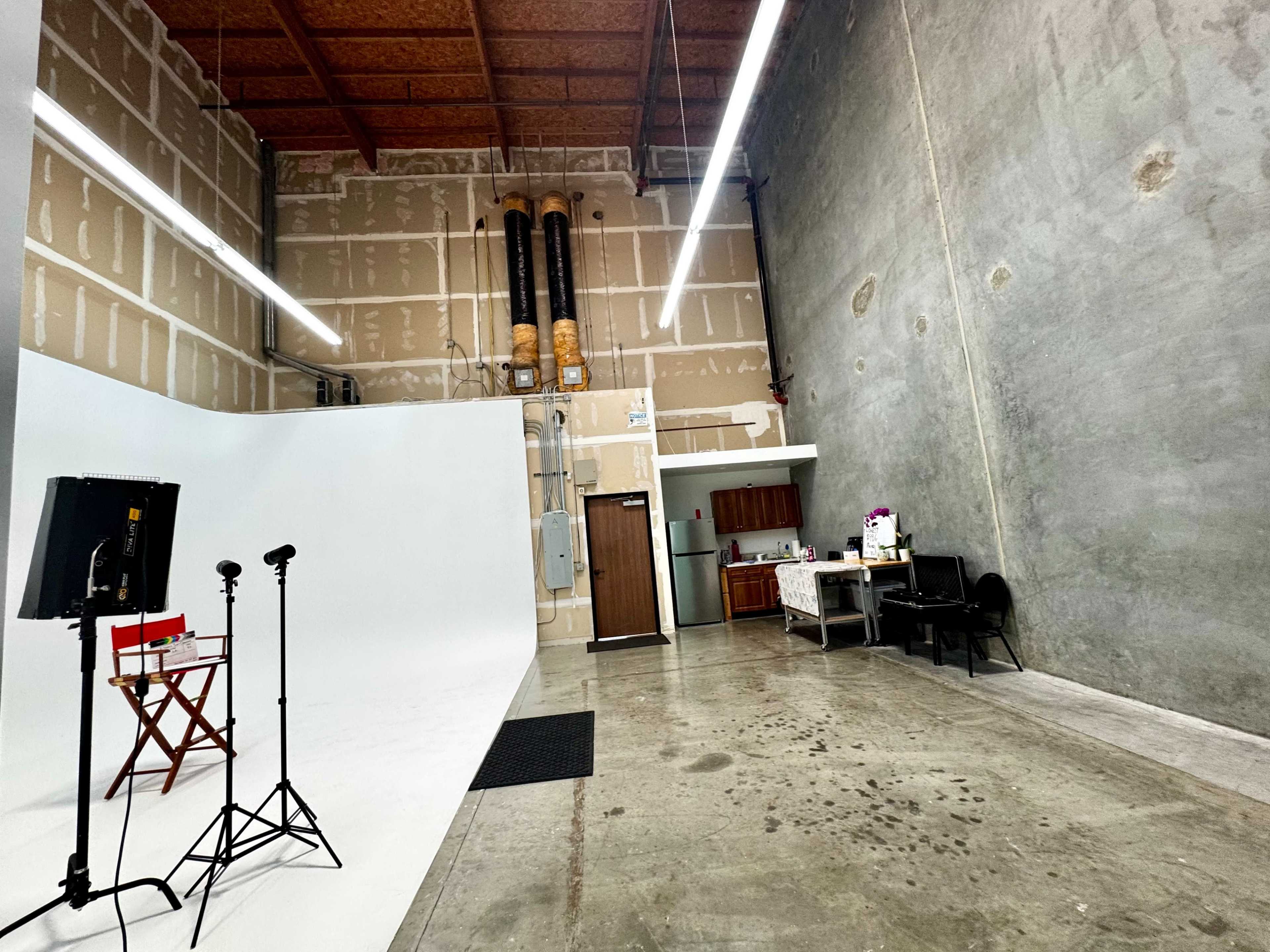 The image shows a spacious studio with a white backdrop, a director's chair, lighting equipment, and a kitchenette area in the corner.