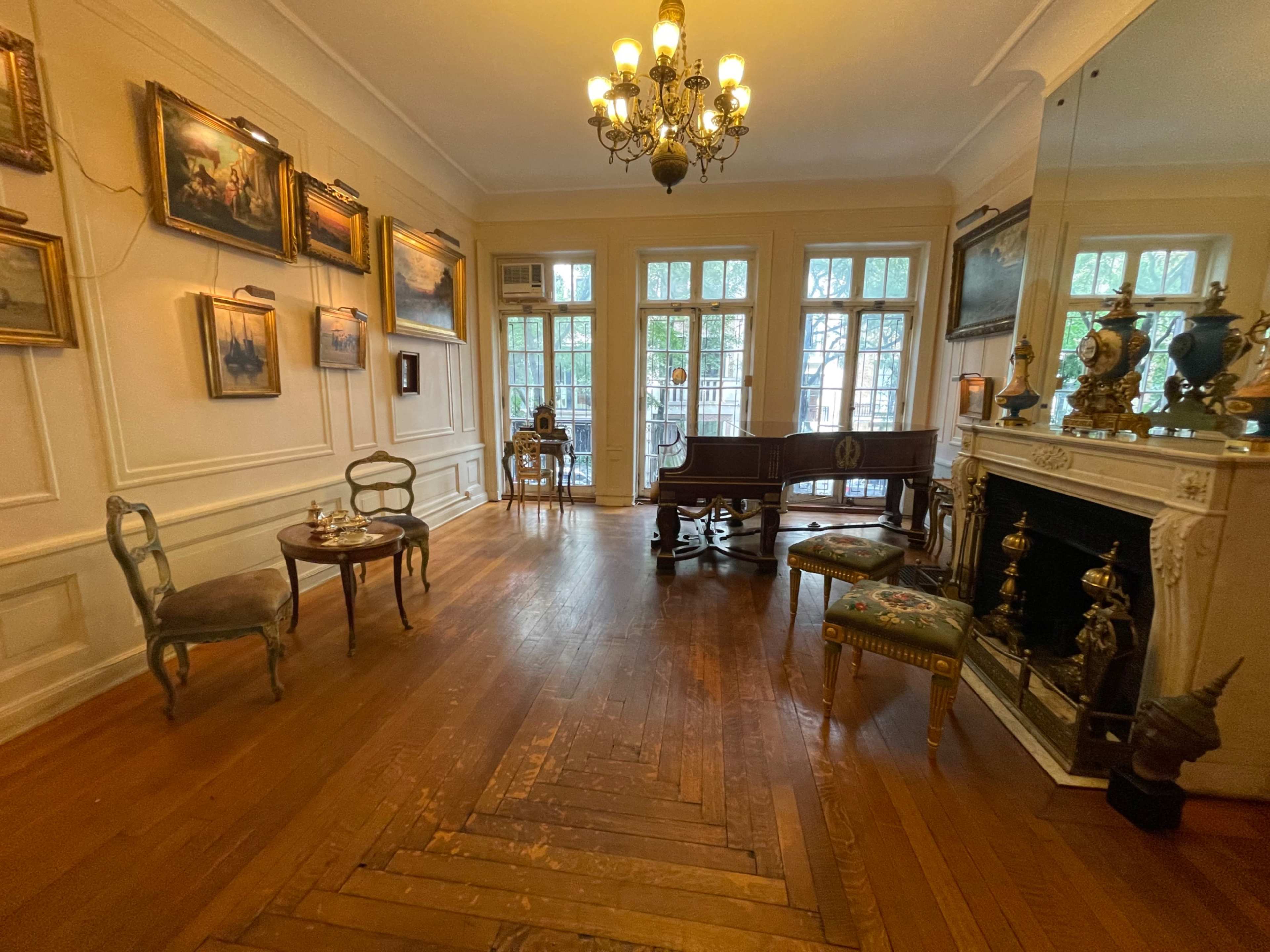 The image shows a room with wooden flooring, multiple paintings on the walls, a fireplace, and antique furniture including a table and chairs.