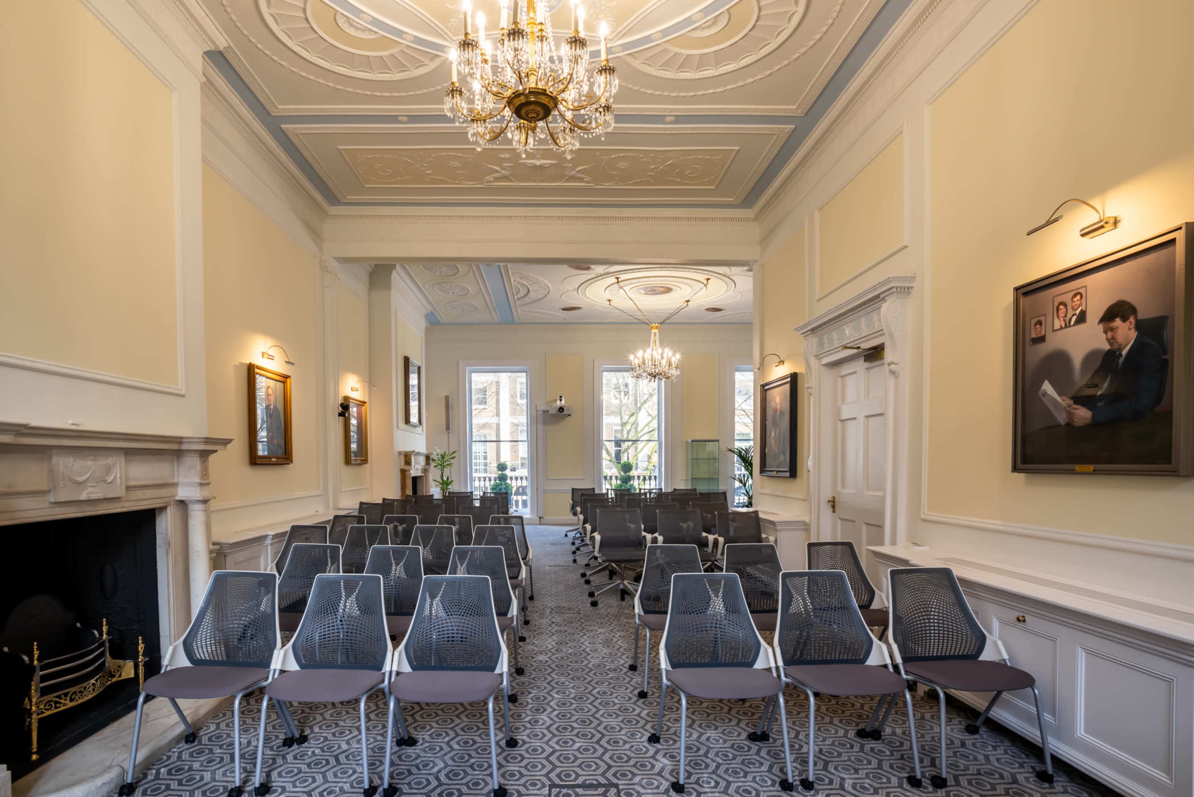 A spacious room with rows of gray mesh chairs facing a fireplace and several framed portraits along the walls.