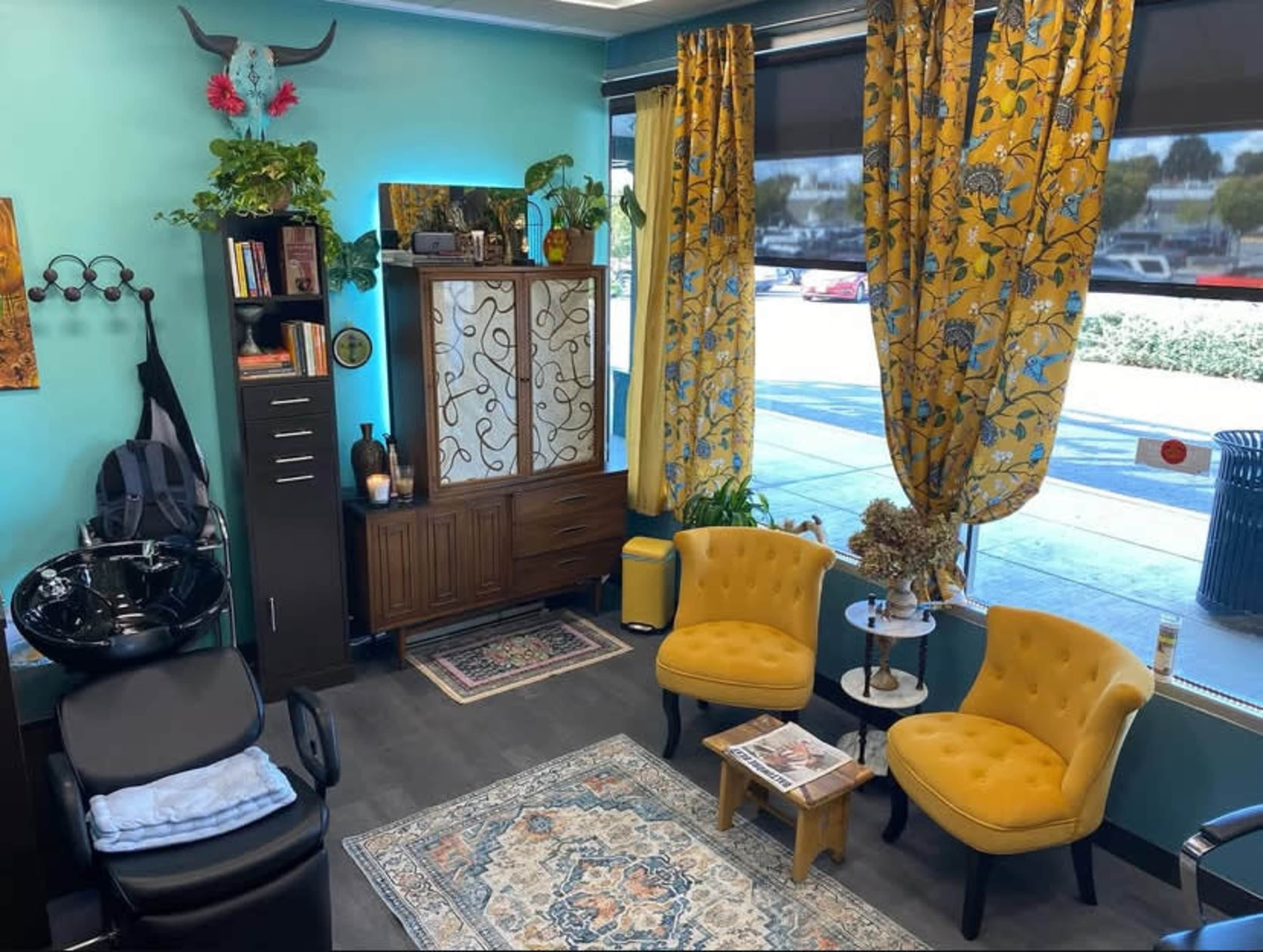 The image shows a bright salon interior featuring two yellow chairs, a cabinet with decorative items, and large windows with patterned curtains.