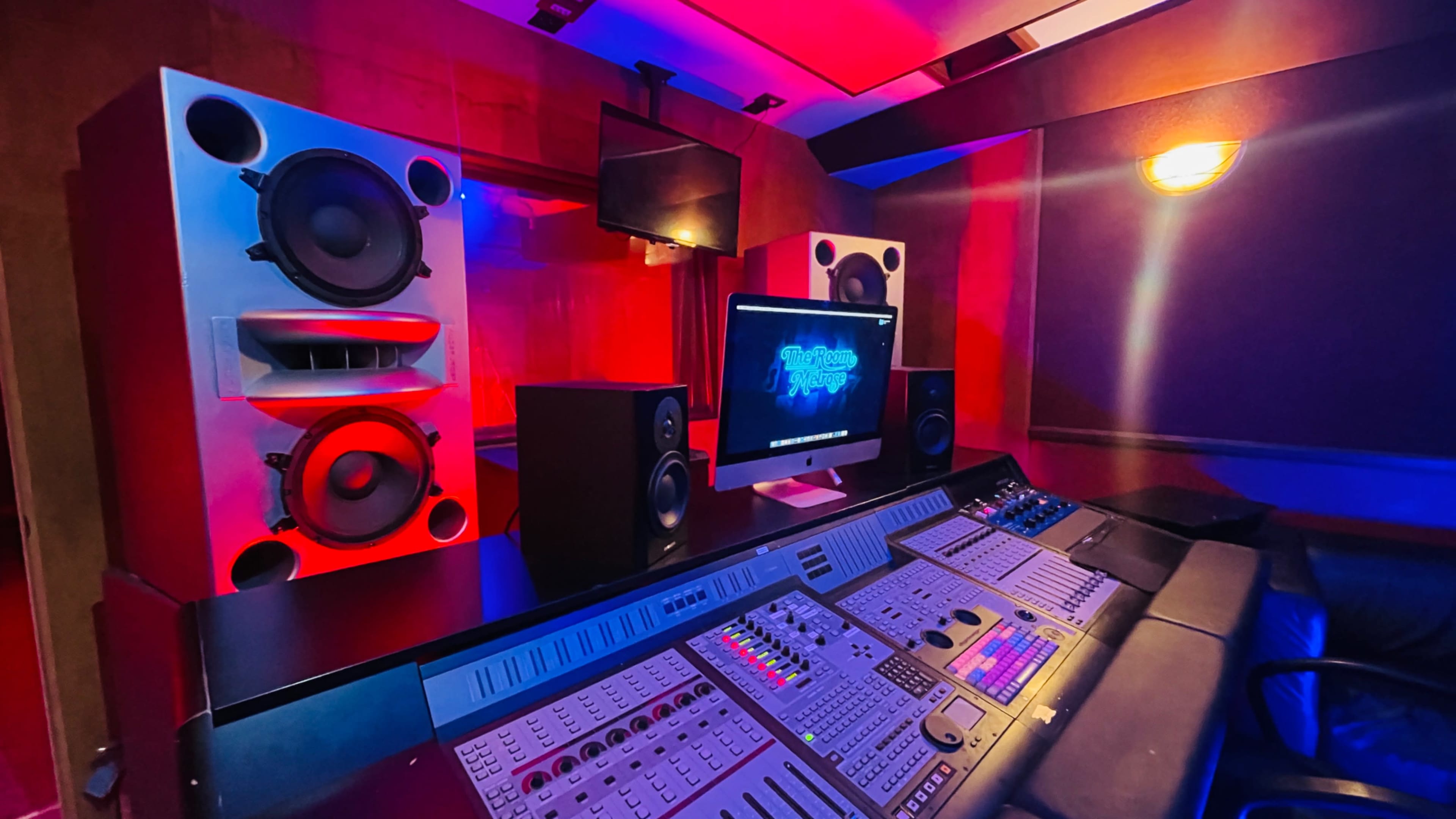 The image shows a dimly lit music studio featuring a mixing console, large speakers, and a computer monitor with audio software displayed.