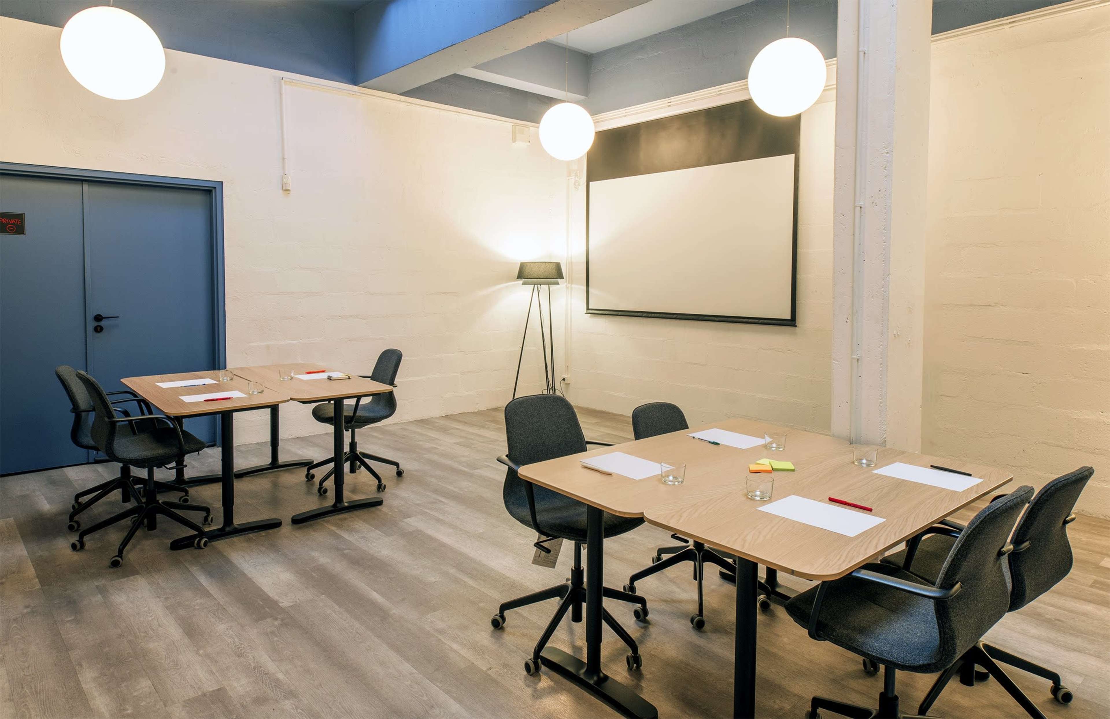 A bright, modern meeting room with two tables arranged for a discussion, each equipped with notepads, pens, and glasses of water, alongside a projector screen at the front.