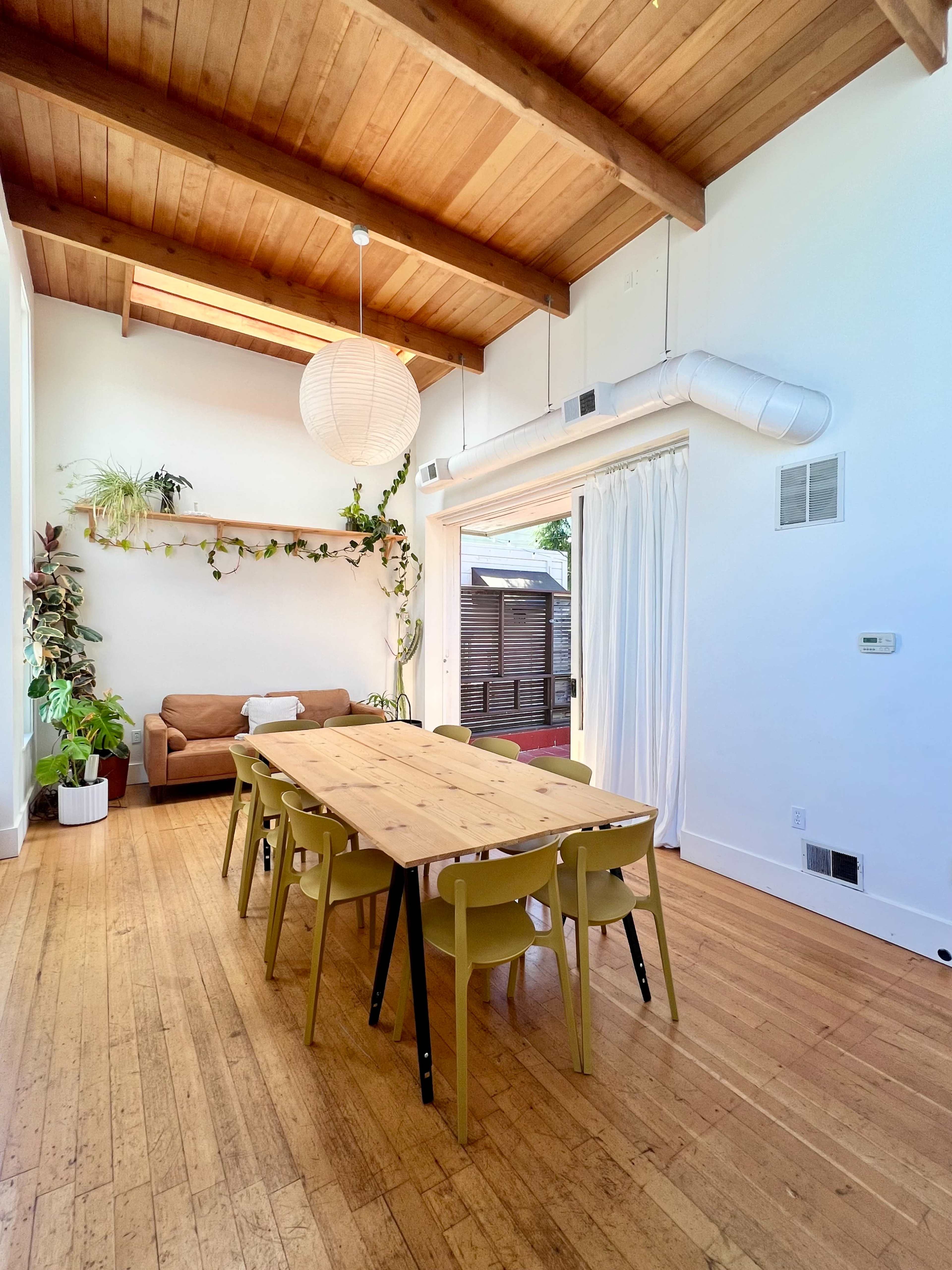 A spacious dining area features a long wooden table with yellow chairs, surrounded by plants and a sofa, under wooden beams and a large pendant light.