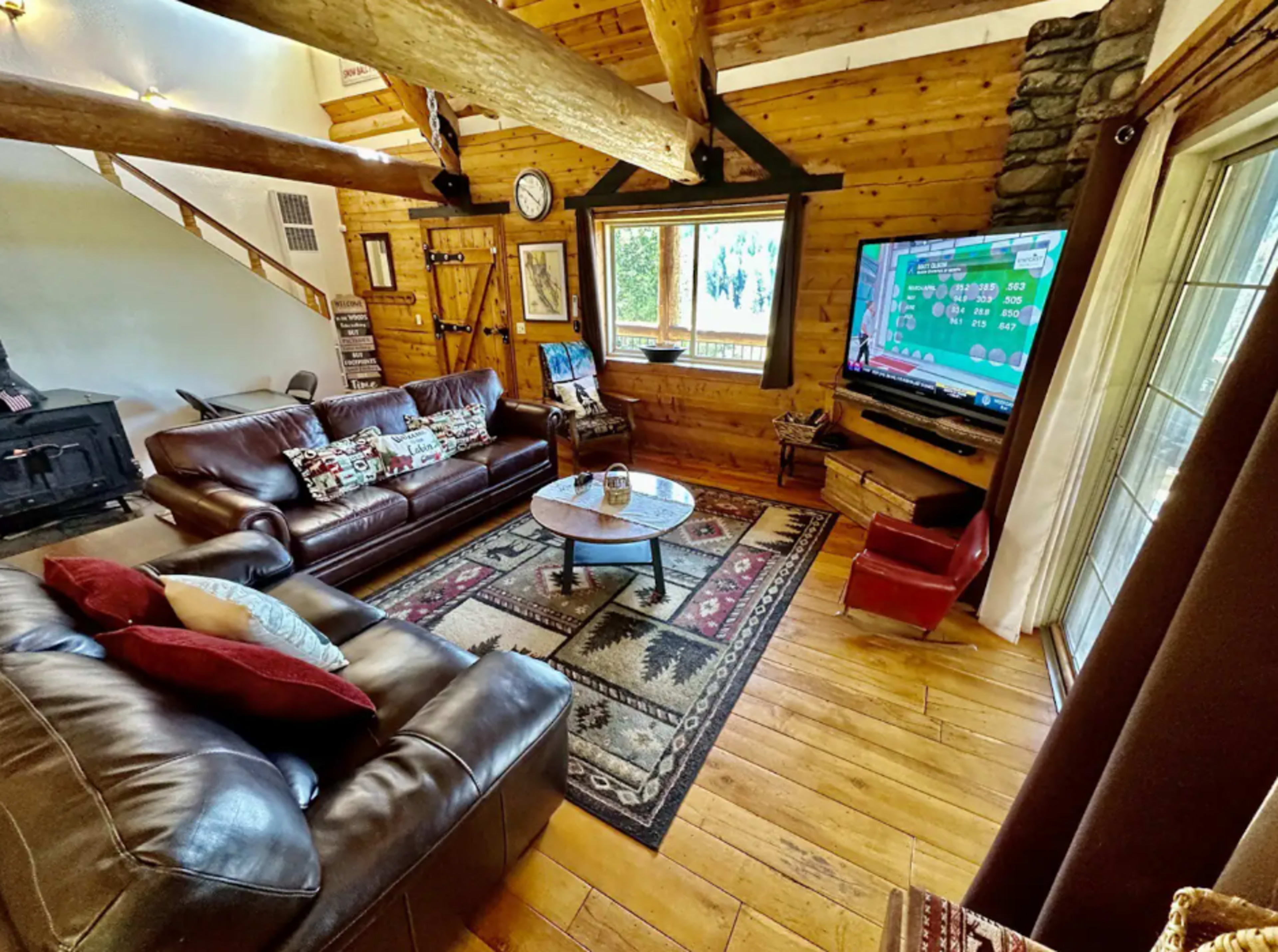 A cozy living room with wooden walls, a stone fireplace, and leather sofas arranged around a circular coffee table.