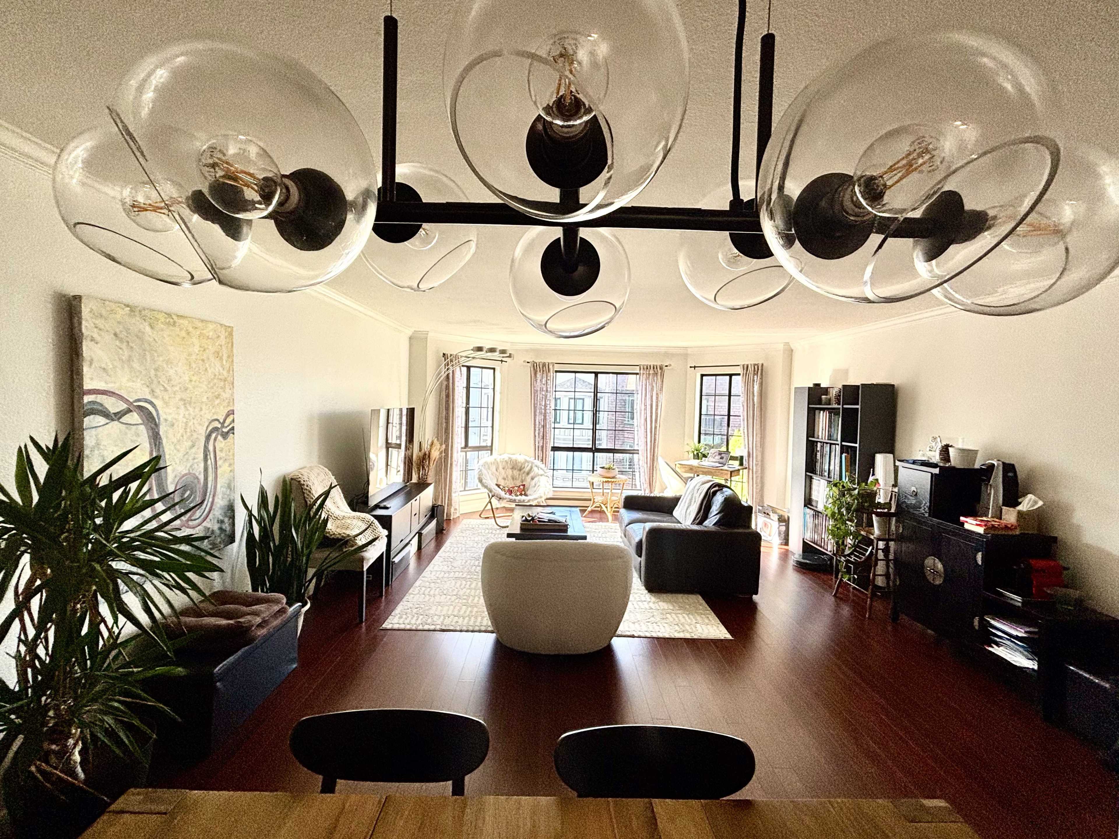 The image shows a spacious living area with wooden flooring, featuring a large sofa, a coffee table, and several windows letting in natural light.