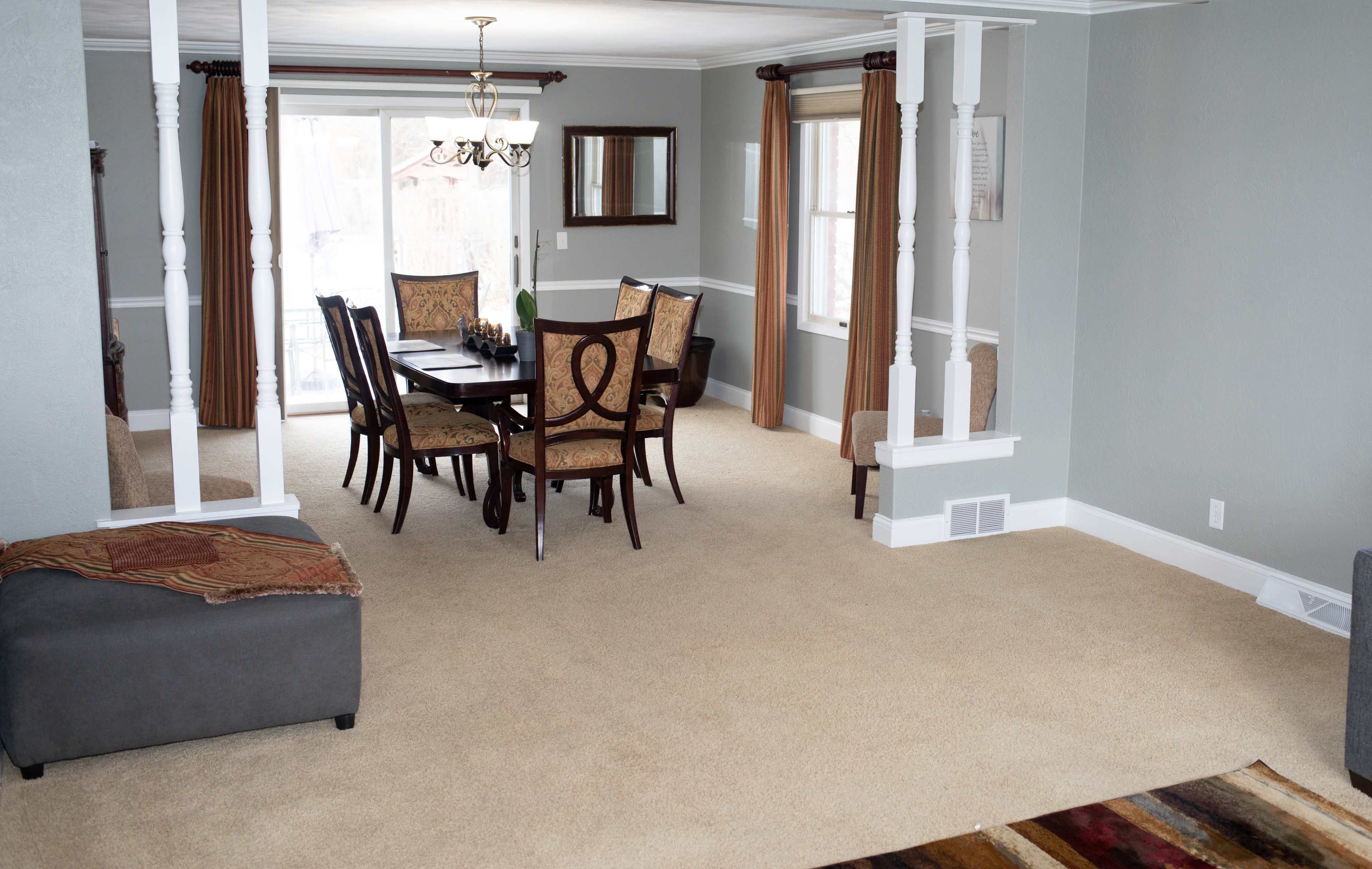The image shows a spacious living and dining area featuring a wooden dining table with chairs, a chandelier, and a nearby gray couch on carpeted flooring.