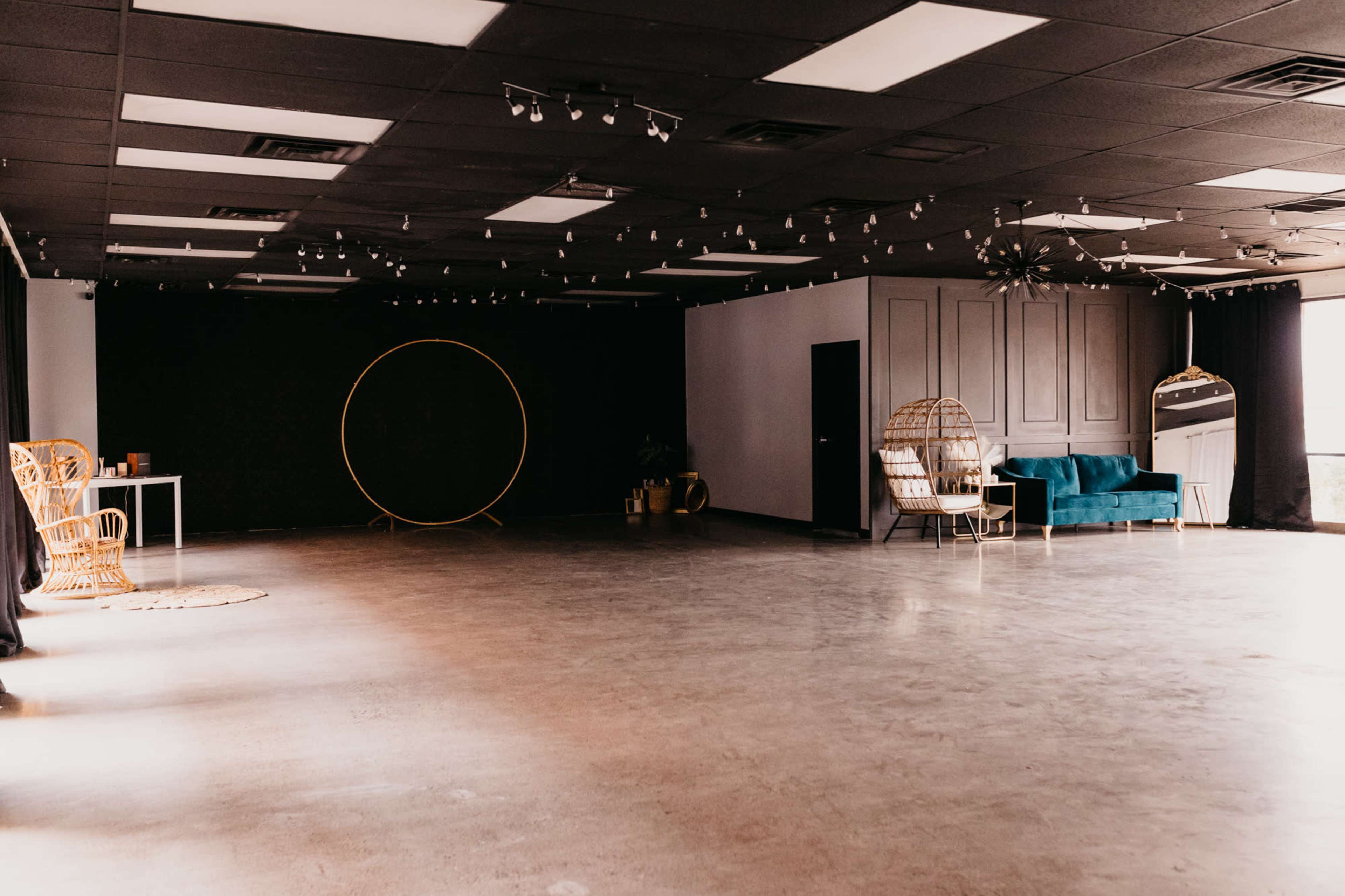 An empty indoor space featuring concrete flooring, a dark wall, a round backdrop, and minimal furnishings including a blue sofa and a table.