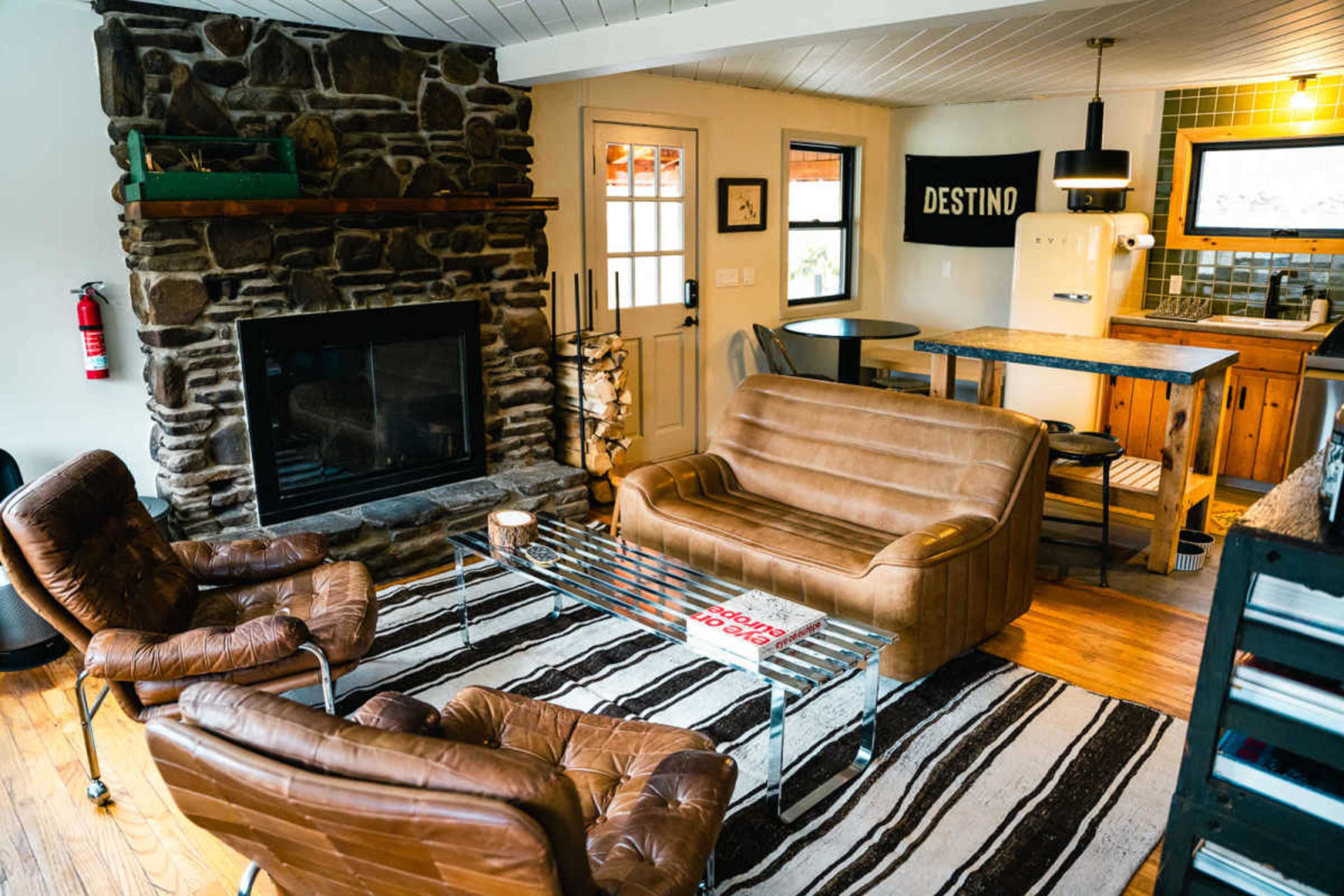 The image shows a modern living area featuring a stone fireplace, brown leather seating, and a striped rug, with a kitchen area visible in the background.