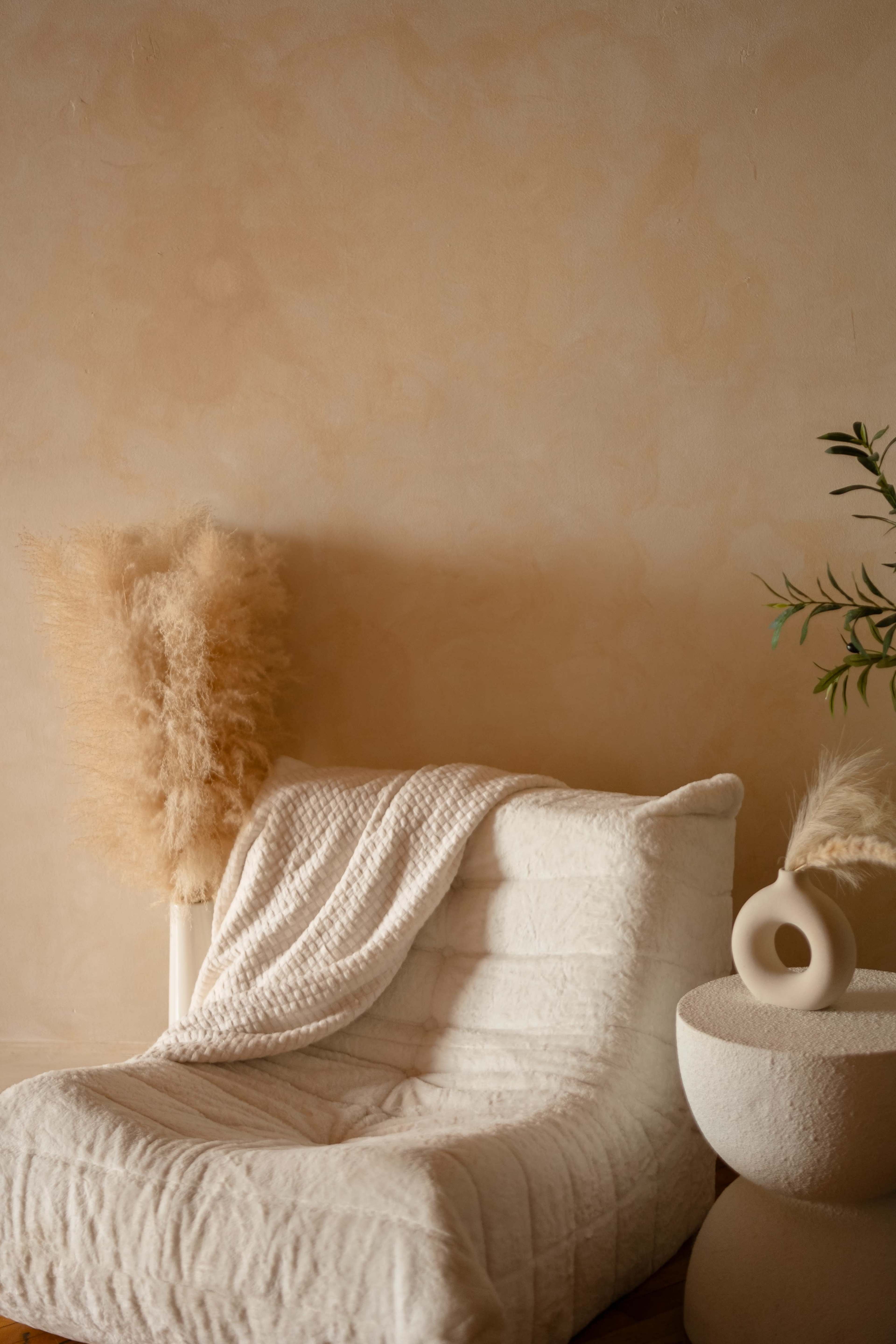 A cream-colored lounge chair with a textured blanket rests beside a round, sculptural table and a cluster of dried pampas grass.