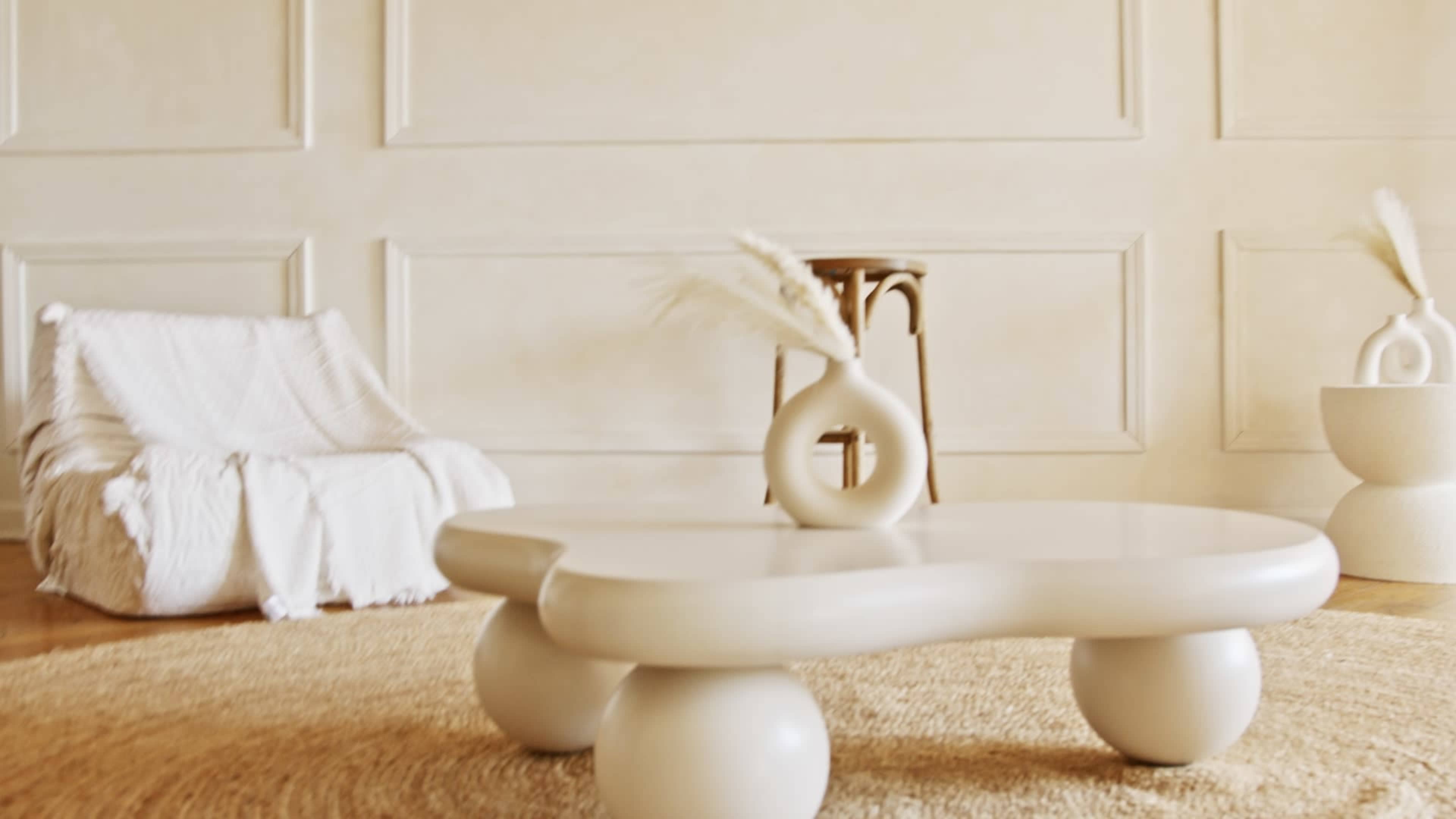 The image features a minimalist living space with a round coffee table, a vase, a textured armchair, and a woven rug on a wooden floor.