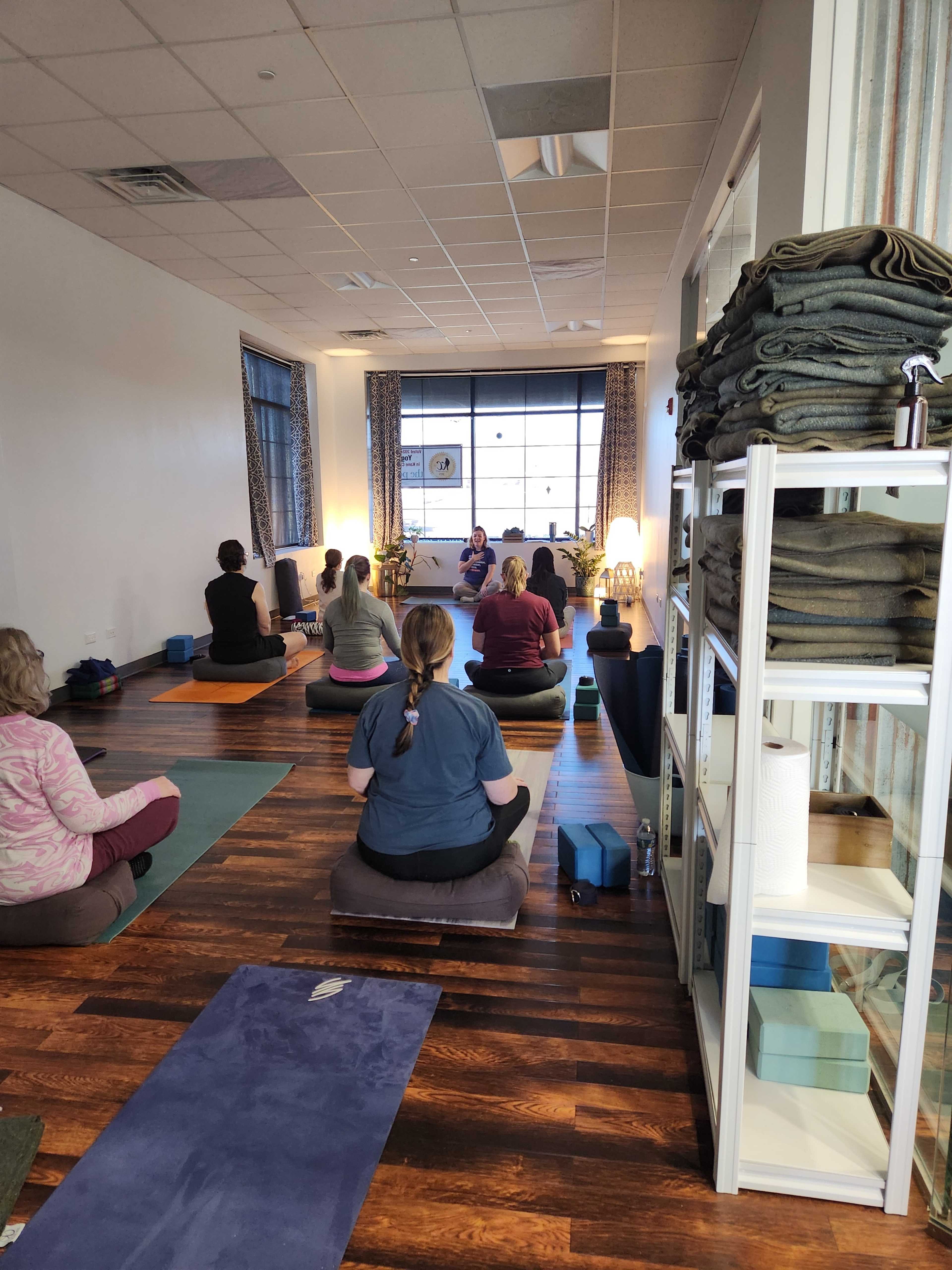 A group of individuals participates in a yoga class in a spacious studio with large windows and neatly organized yoga mats.