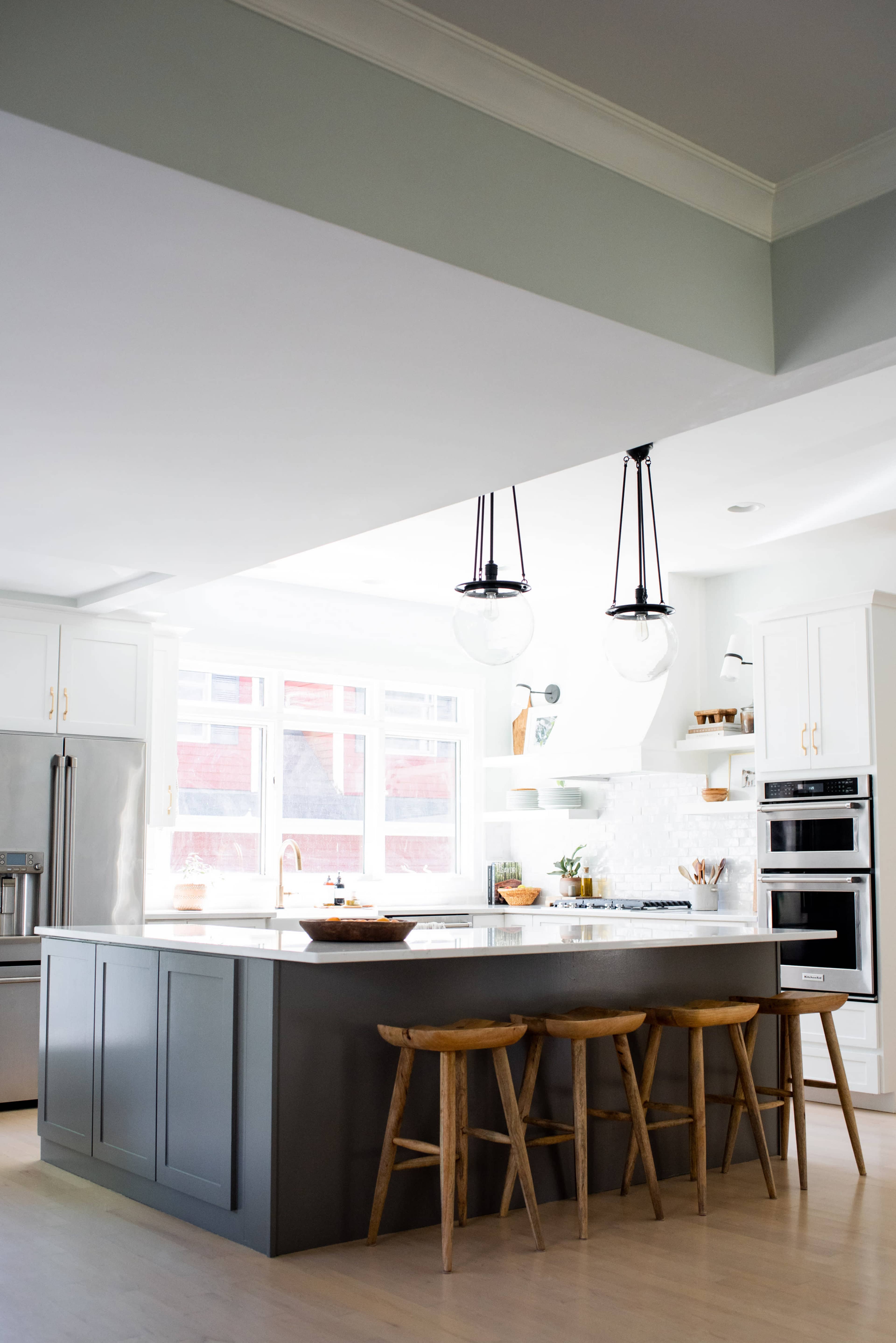 The image shows a modern kitchen featuring an island with wooden stools, stainless steel appliances, and large windows that allow natural light to illuminate the space.