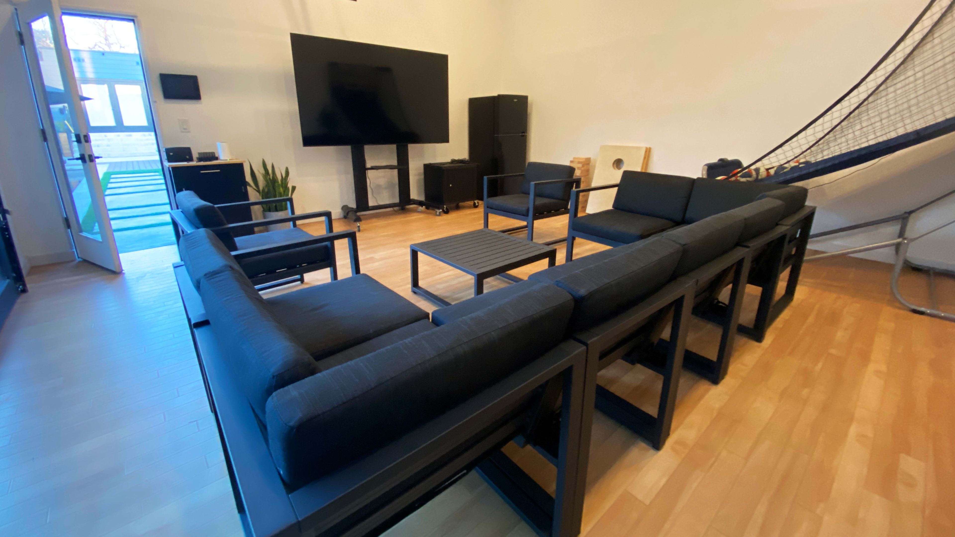 A modern living room with several black sofas arranged around a coffee table, a large TV on the wall, and a staircase leading to an upper level.