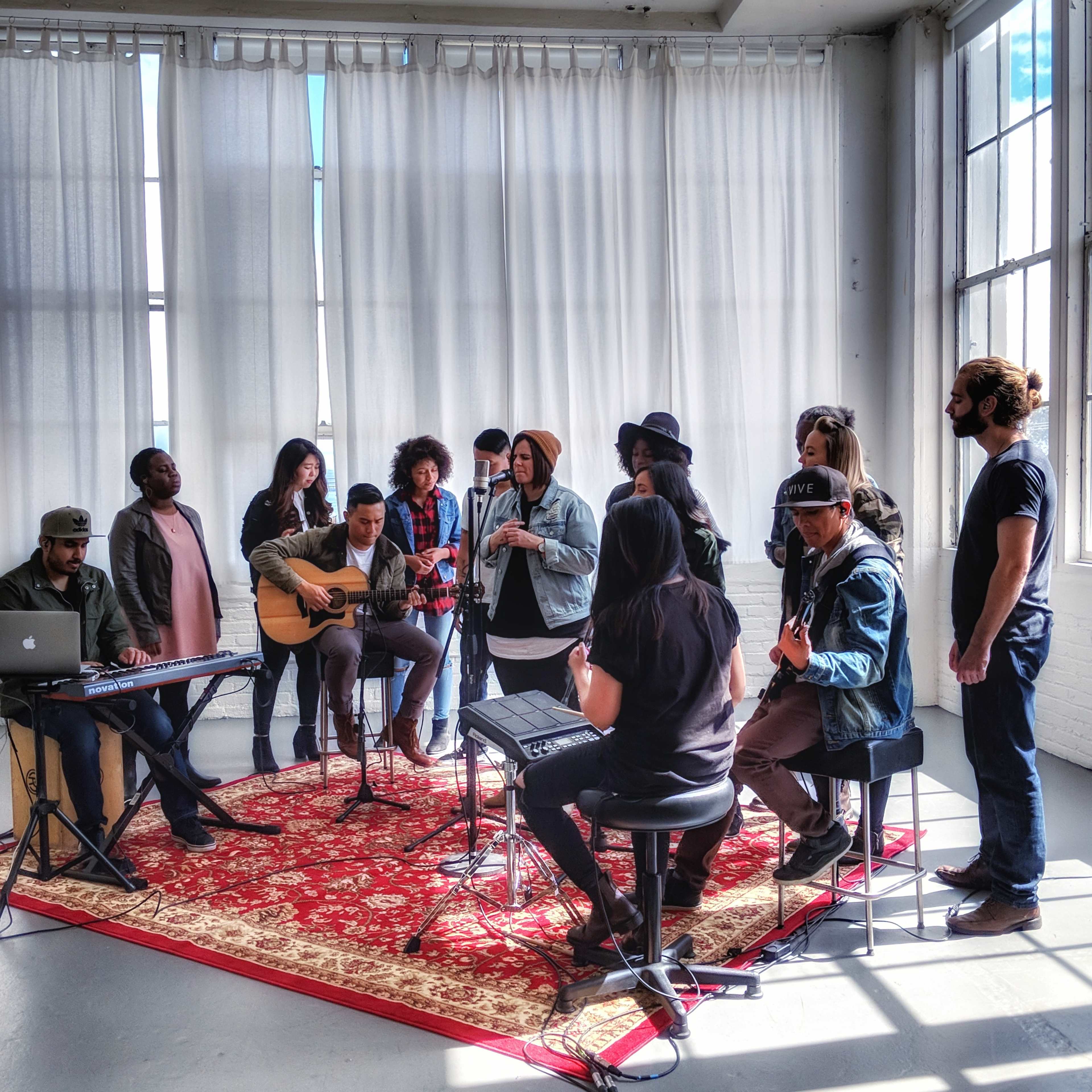 A group of musicians performs together in a sunlit room with large windows and white curtains, surrounded by a red patterned rug.