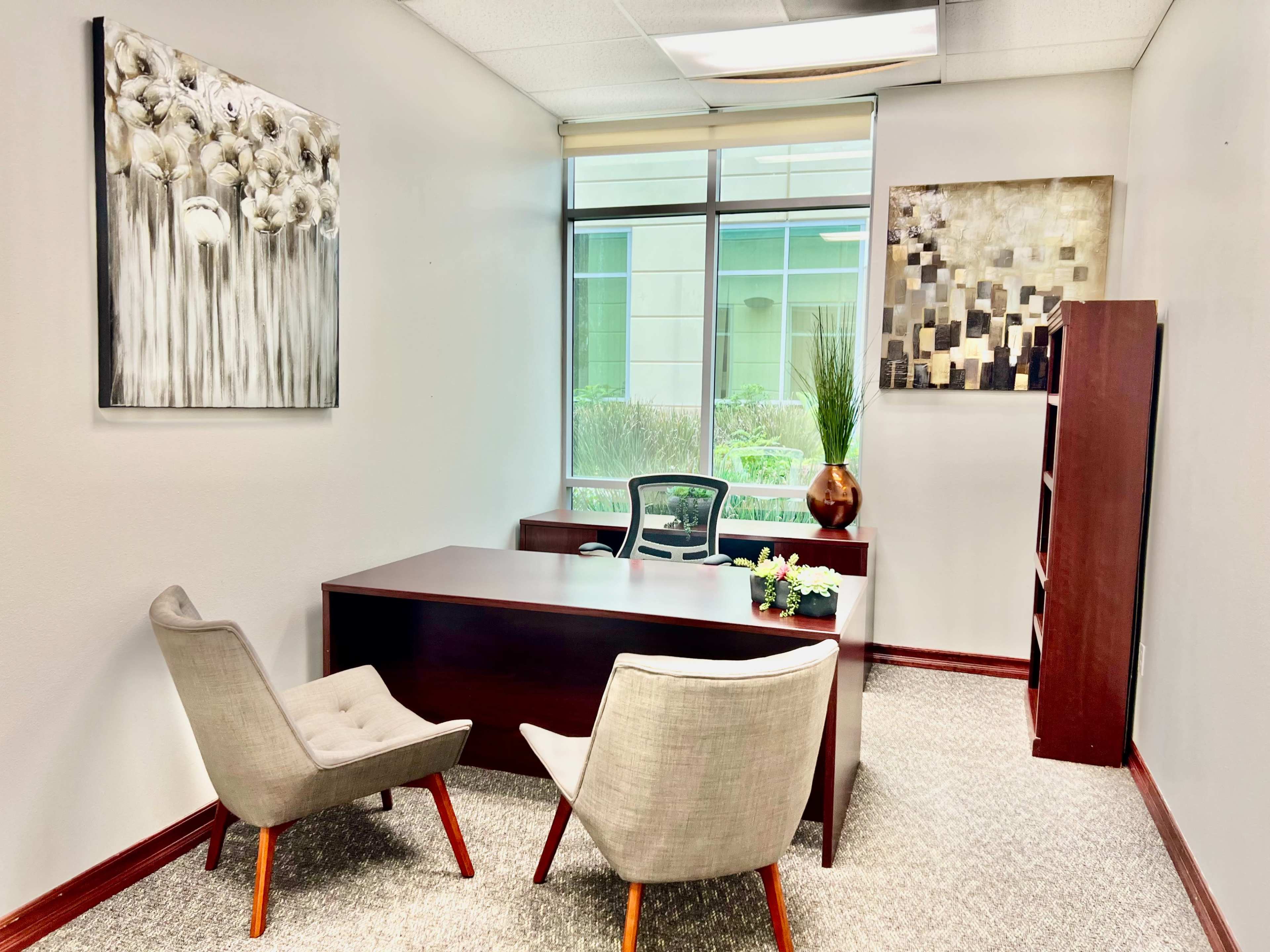 The image shows a modern office space featuring a wooden desk, two upholstered chairs, wall art, and a window with plants outside.
